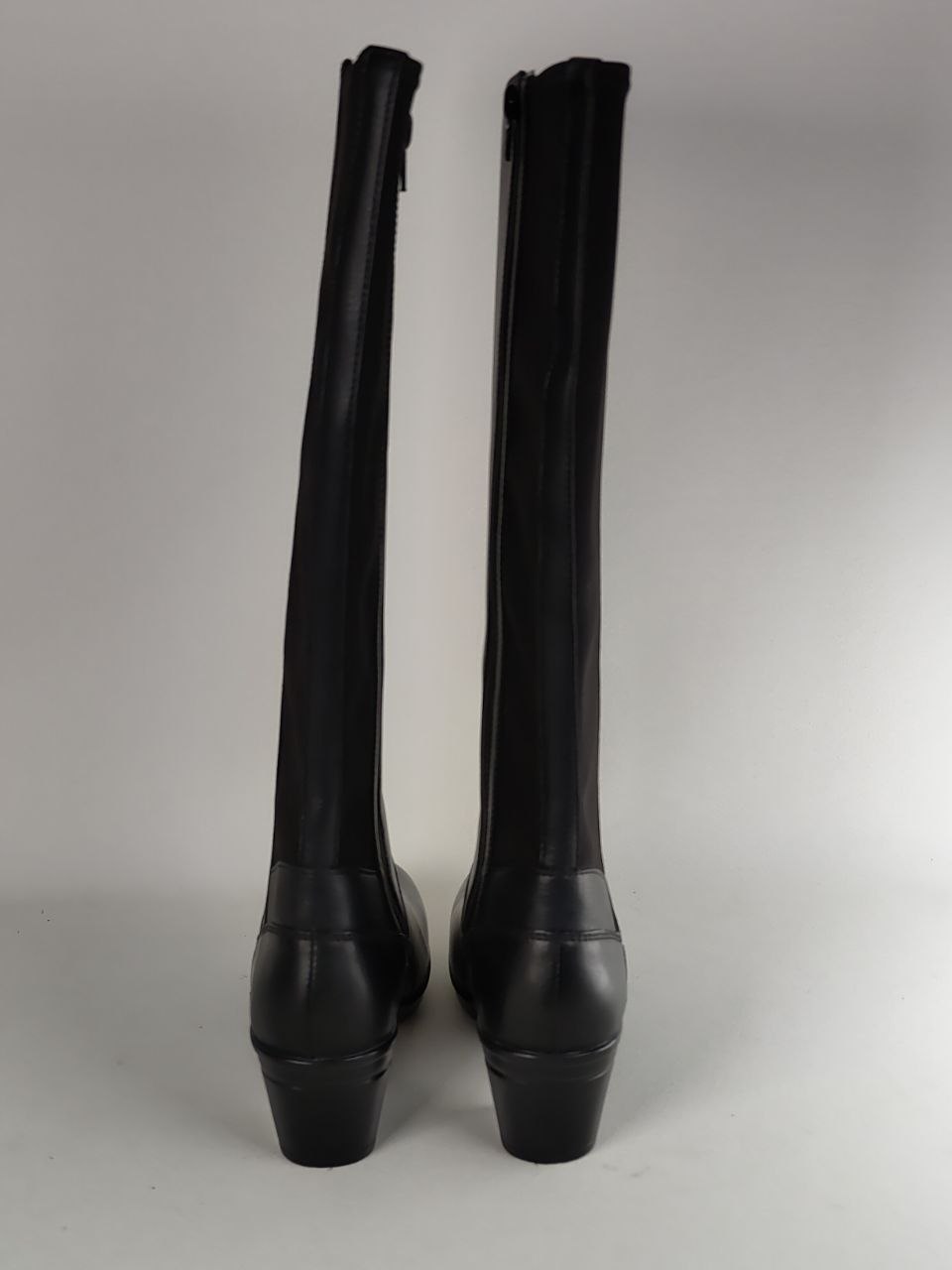 A pair of black leather knee-high boots with block heels, shown from the back against a neutral gray background.
