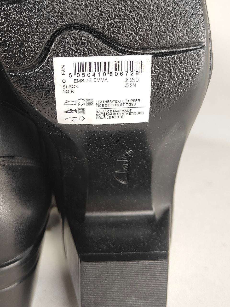 Close-up of the underside of a black leather shoe, showing a white size and material label with barcode and text, plus the embossed Clarks logo on the heel.