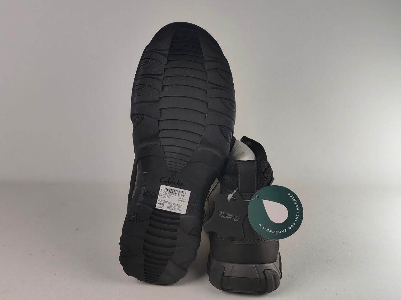 A pair of black outdoor boots shown from the back, with one boot standing upright to display the textured black sole and the other lying down to show the heel and side. A green circular hangtag is attached, and a white size label is visible on the sole.