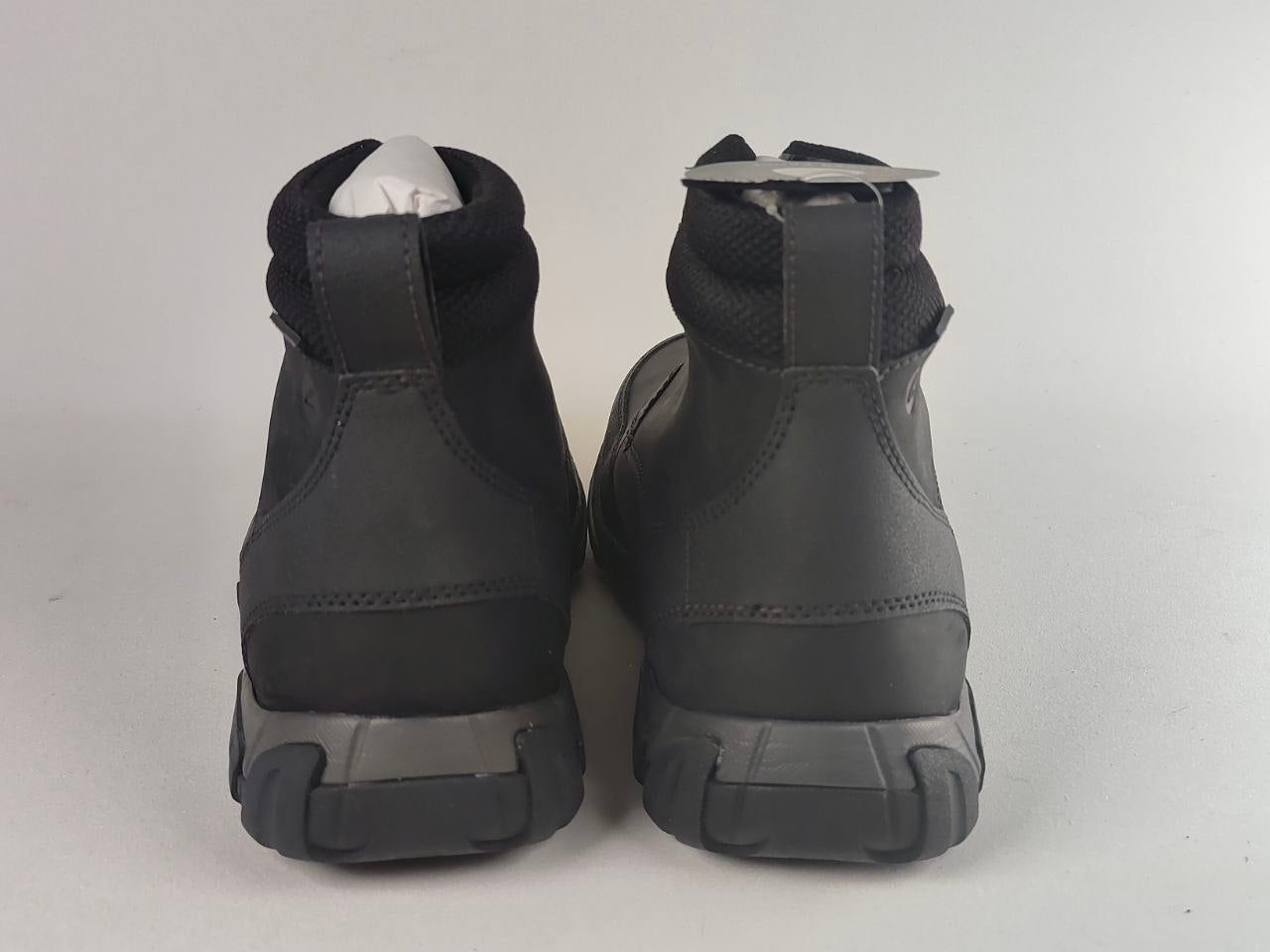 A pair of black high-top boots shown from the back, featuring a matte synthetic upper, padded collar, and chunky black sole, photographed against a light gray background.