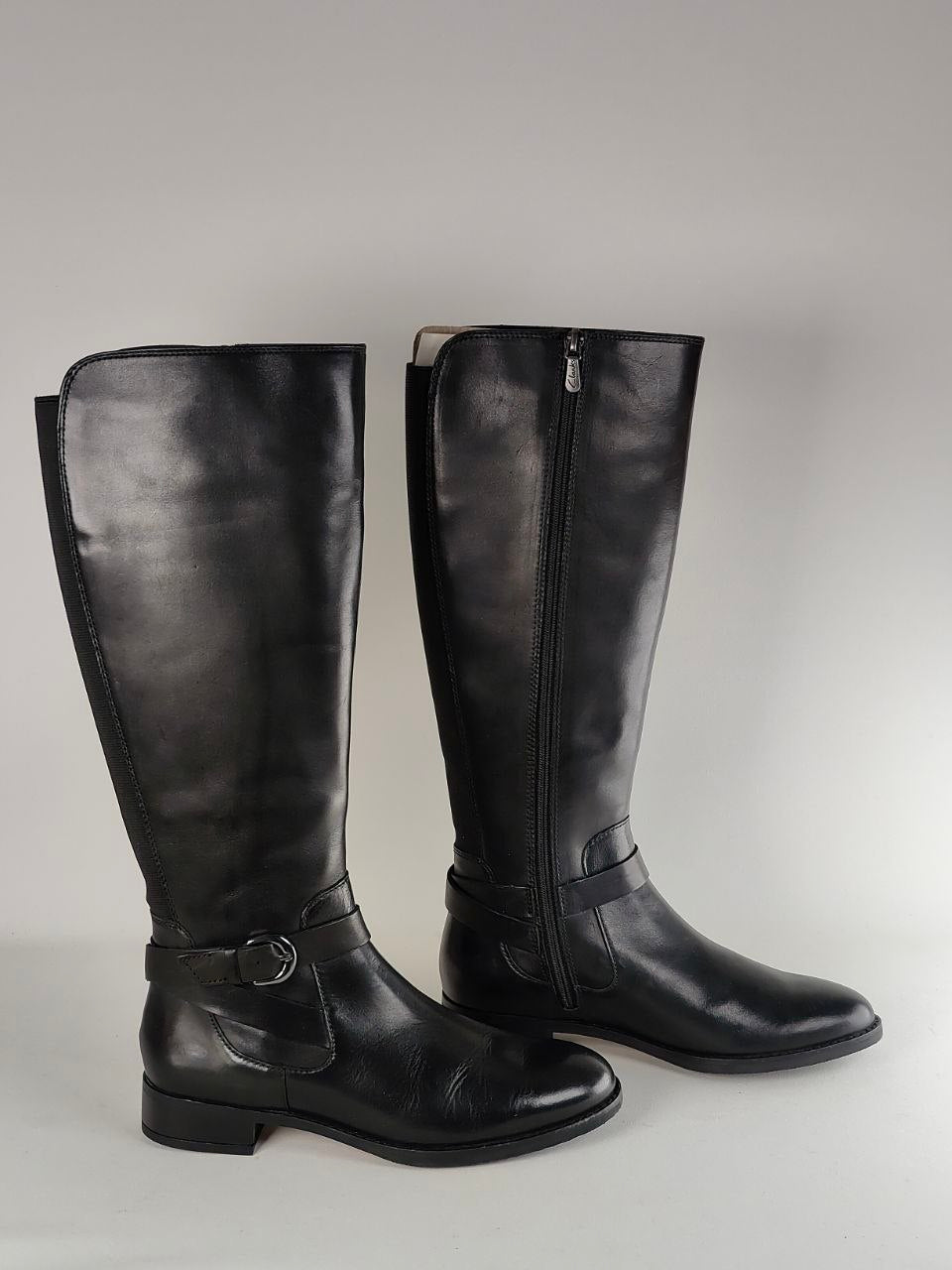 A pair of black leather knee-high riding boots with side zippers and a buckle strap detail on the lower shaft, shown against a neutral gray background.