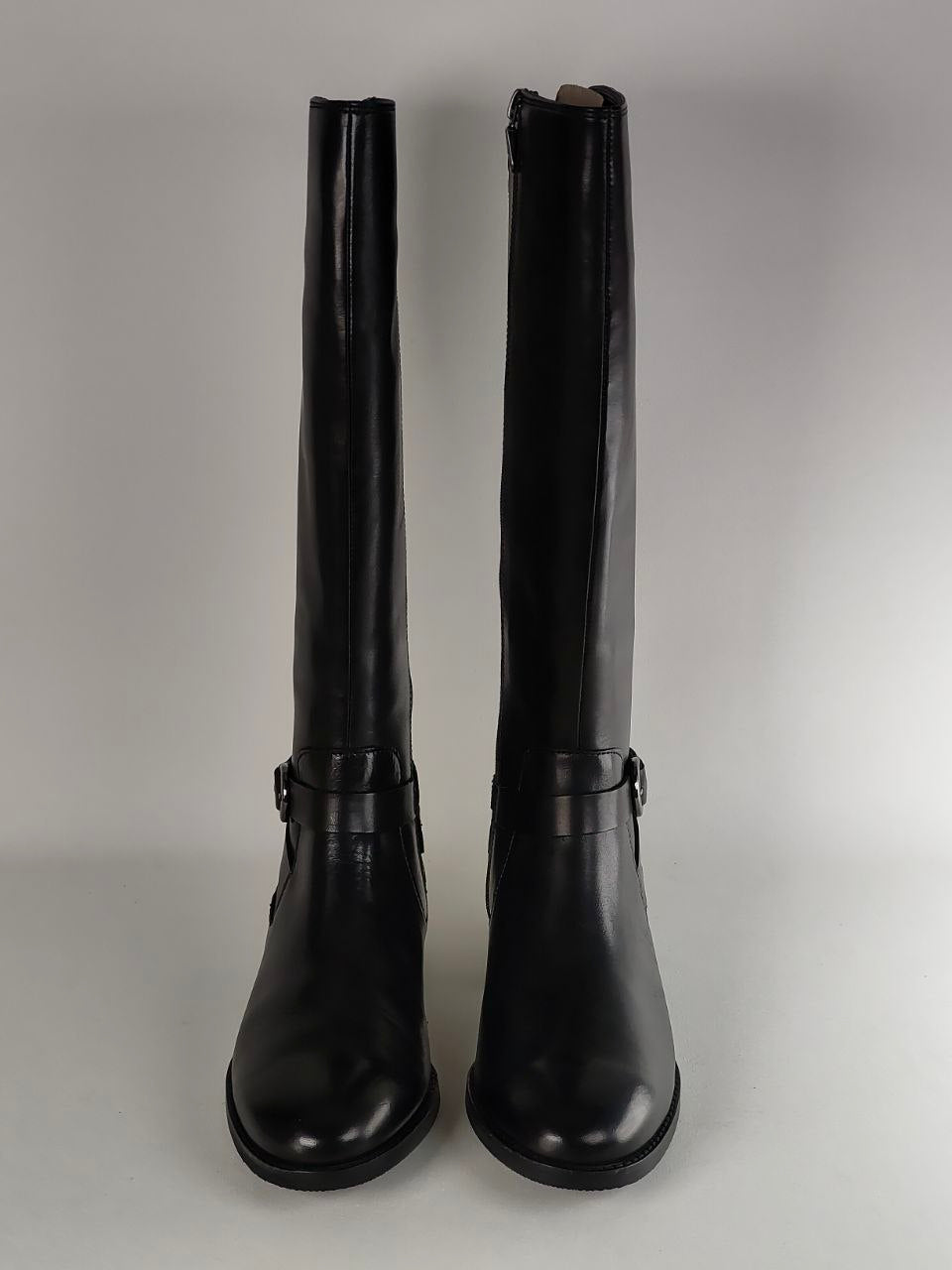 A pair of black leather knee-high riding boots with buckle straps at the lower calf, shown against a neutral gray background.