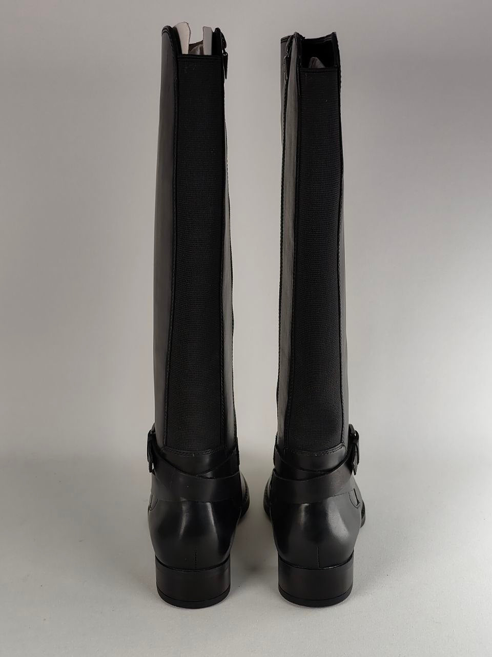A pair of black knee-high riding boots shown from the back, featuring a smooth leather shaft and a stacked block heel, set against a neutral gray background.