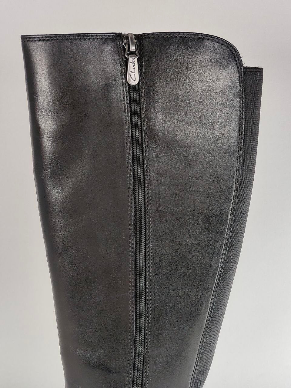 A close-up of a black leather knee-high boot featuring a central metal zipper with a silver pull tab.