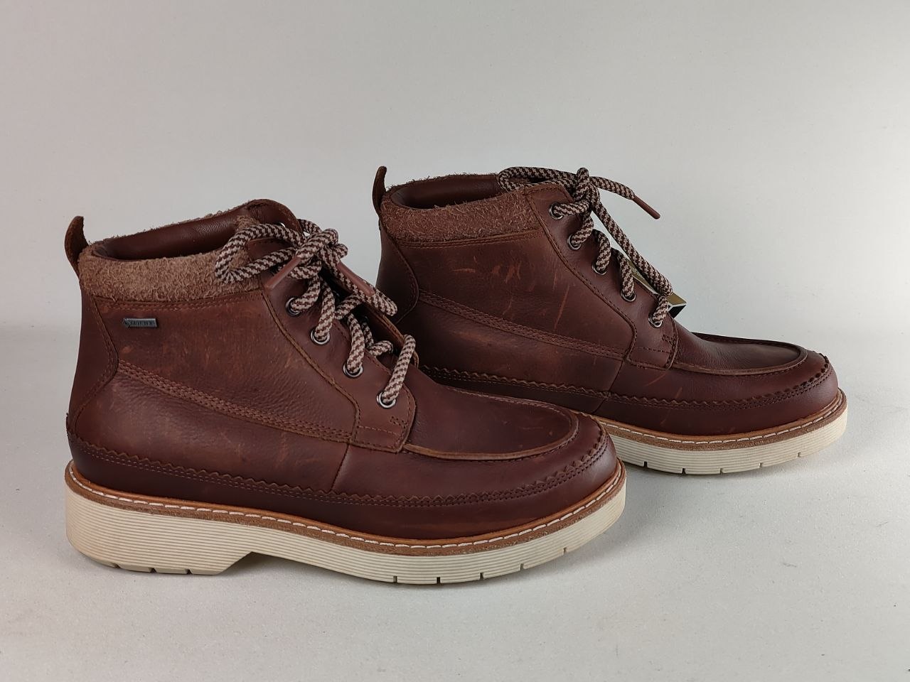 A pair of dark brown leather ankle boots with thick off-white soles and brown-and-white braided laces, shown on a neutral light gray background.