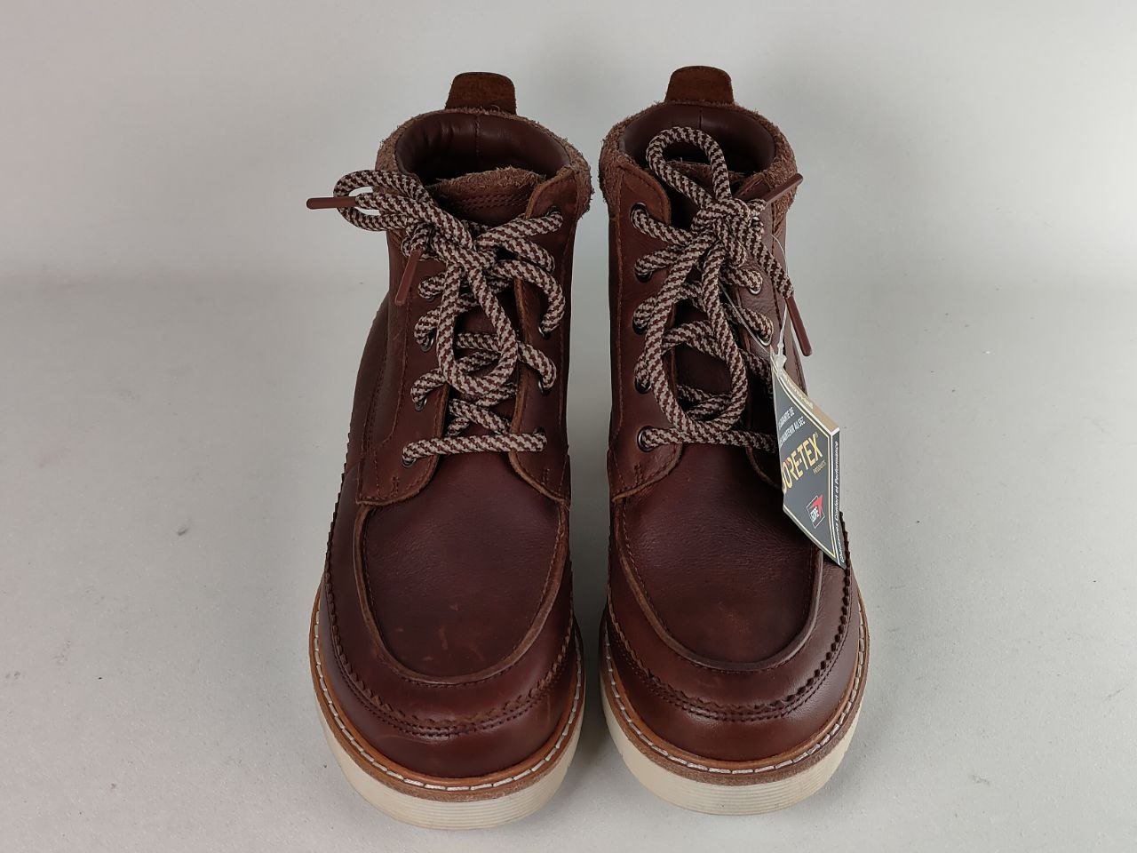 A pair of brown leather lace-up ankle boots with thick white soles and brown-and-white braided laces, shown against a light gray background. A hangtag is attached to the right boot.