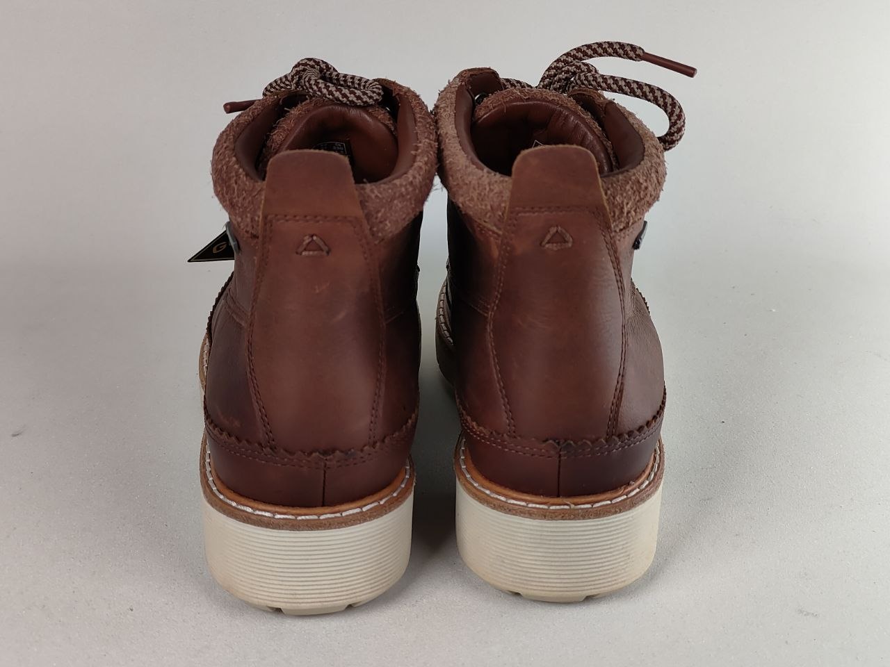 A pair of brown leather ankle boots shown from the back, featuring cream-colored soles and brown laces, set against a plain light gray background.