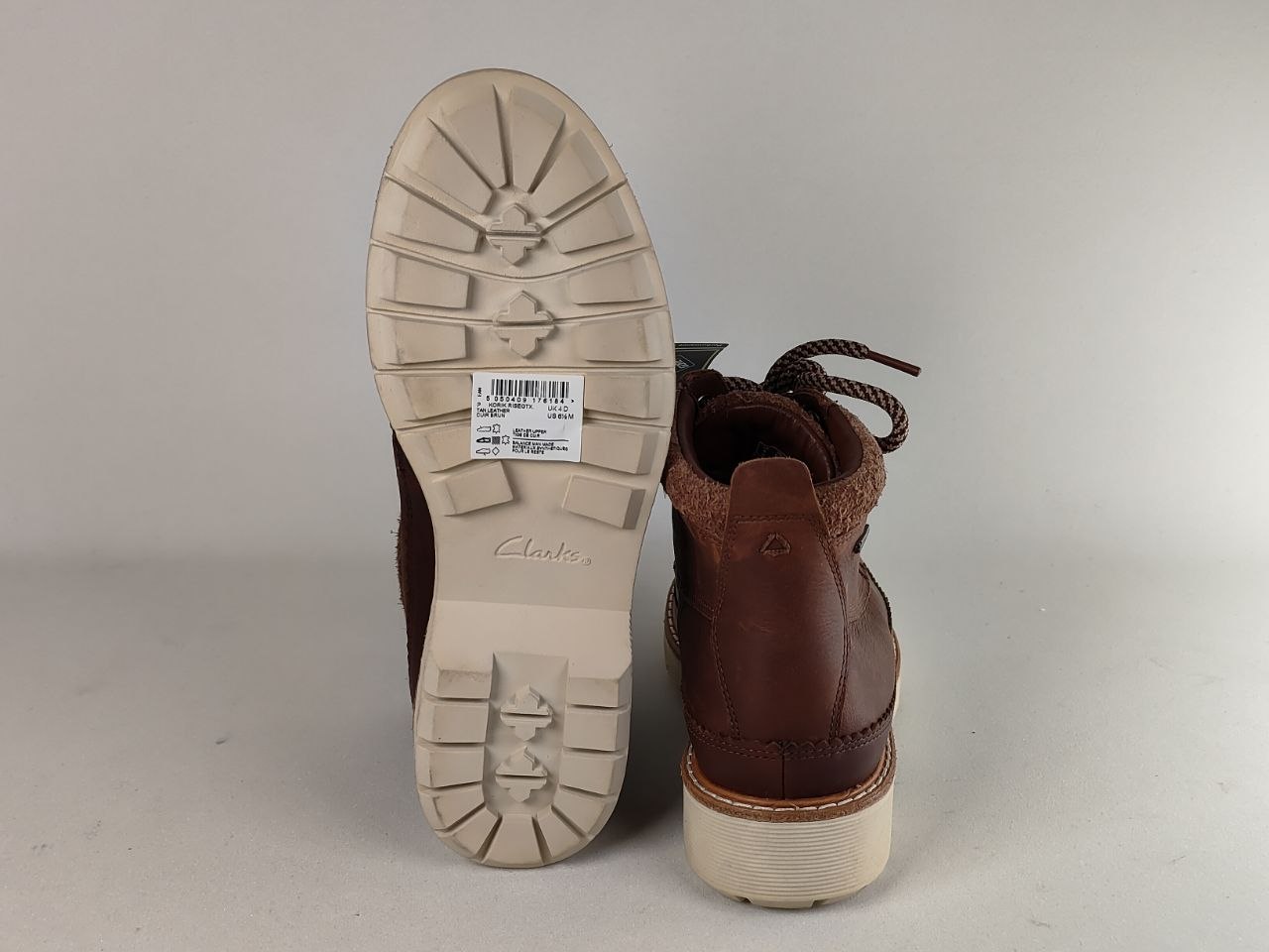 A pair of brown leather ankle boots shown from the back, with one boot standing upright to display the sole and the other lying flat to show the heel and lace area. The soles are off-white, and a Clarks brand tag is visible on the sole.