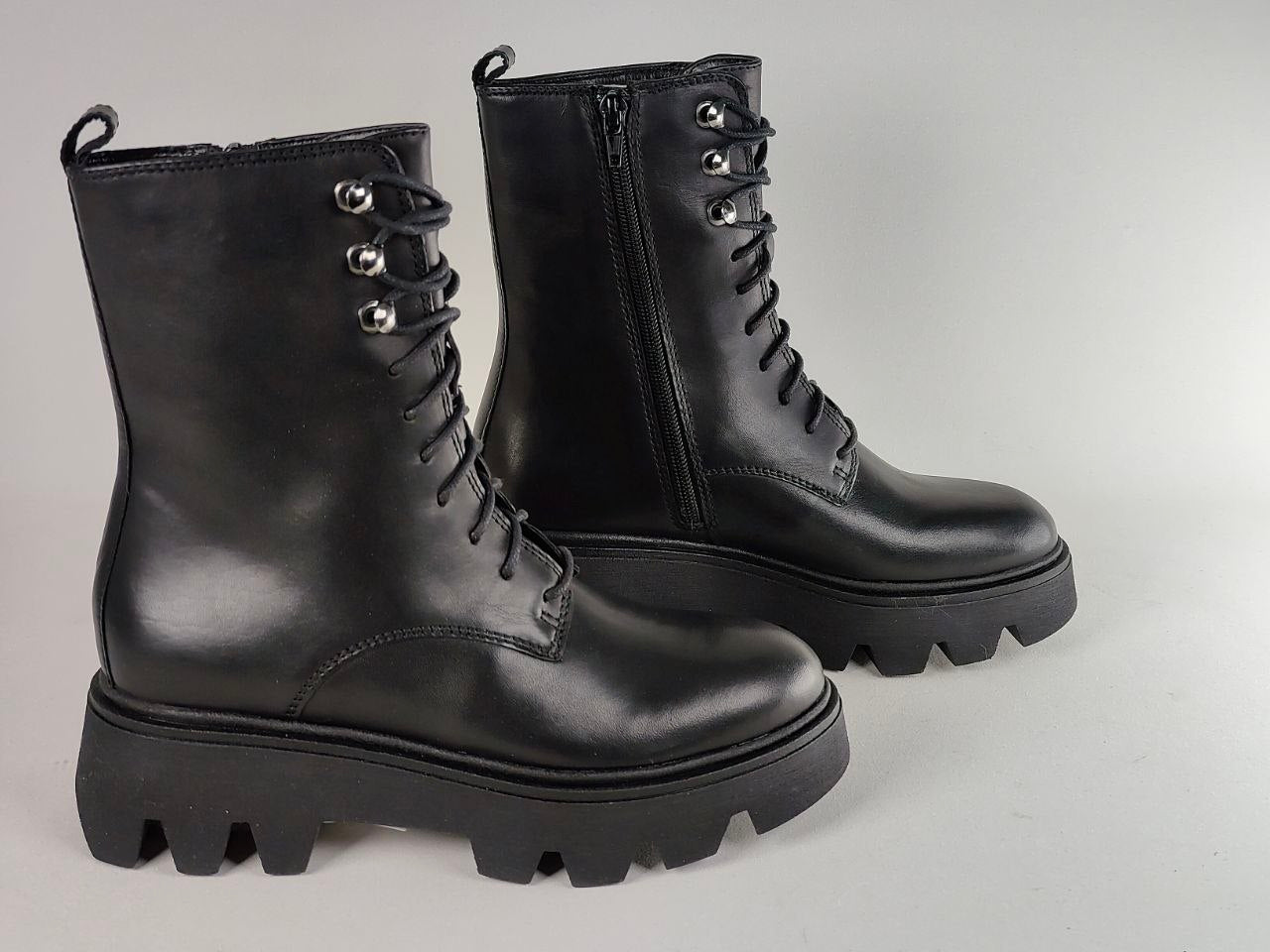A pair of black leather lace-up ankle boots with chunky lug soles and silver-tone eyelets, photographed on a light gray studio background.