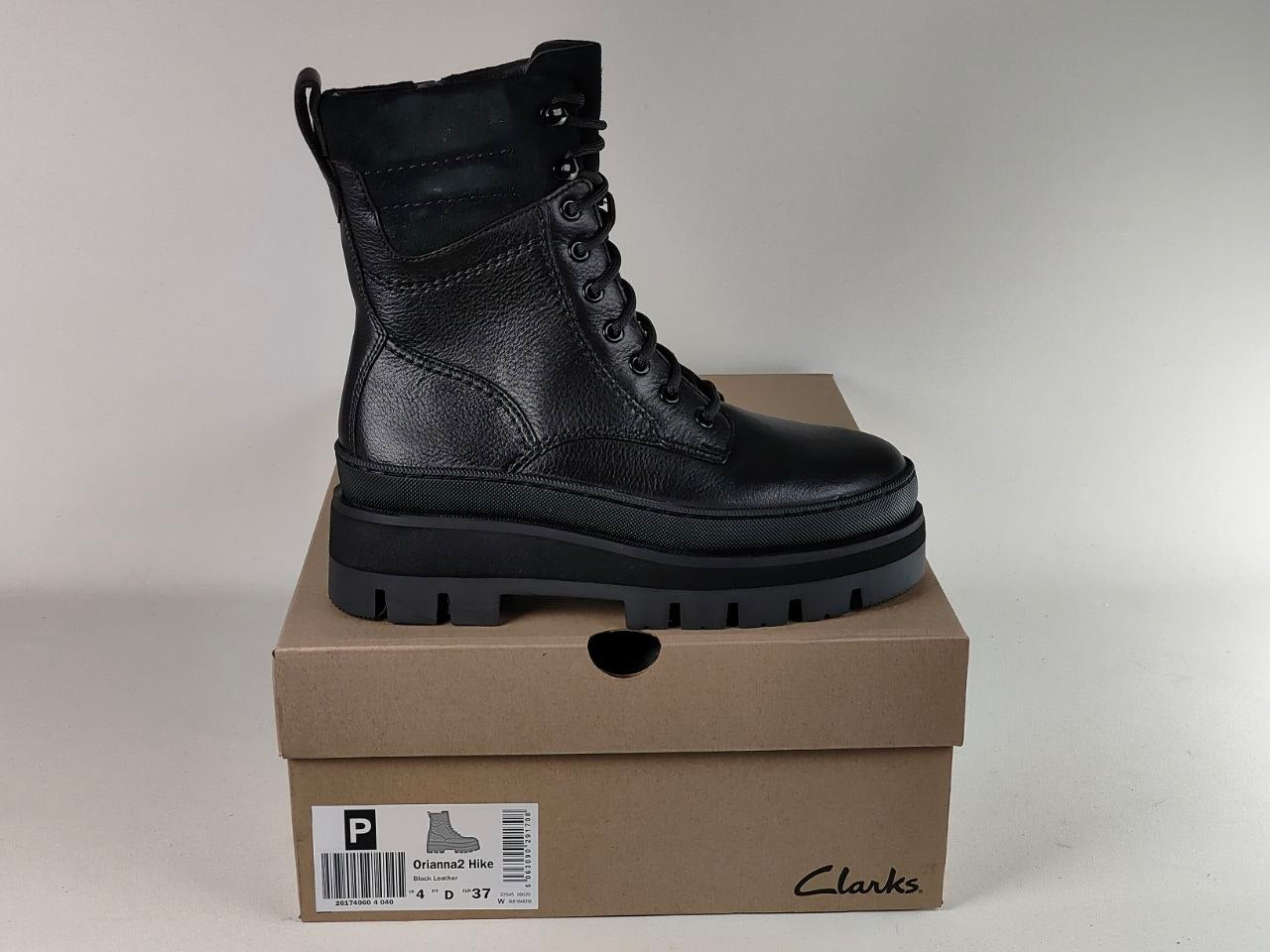 A black leather Clarks ankle boot with a chunky lug sole, displayed on top of its brown cardboard shoebox against a light gray background.