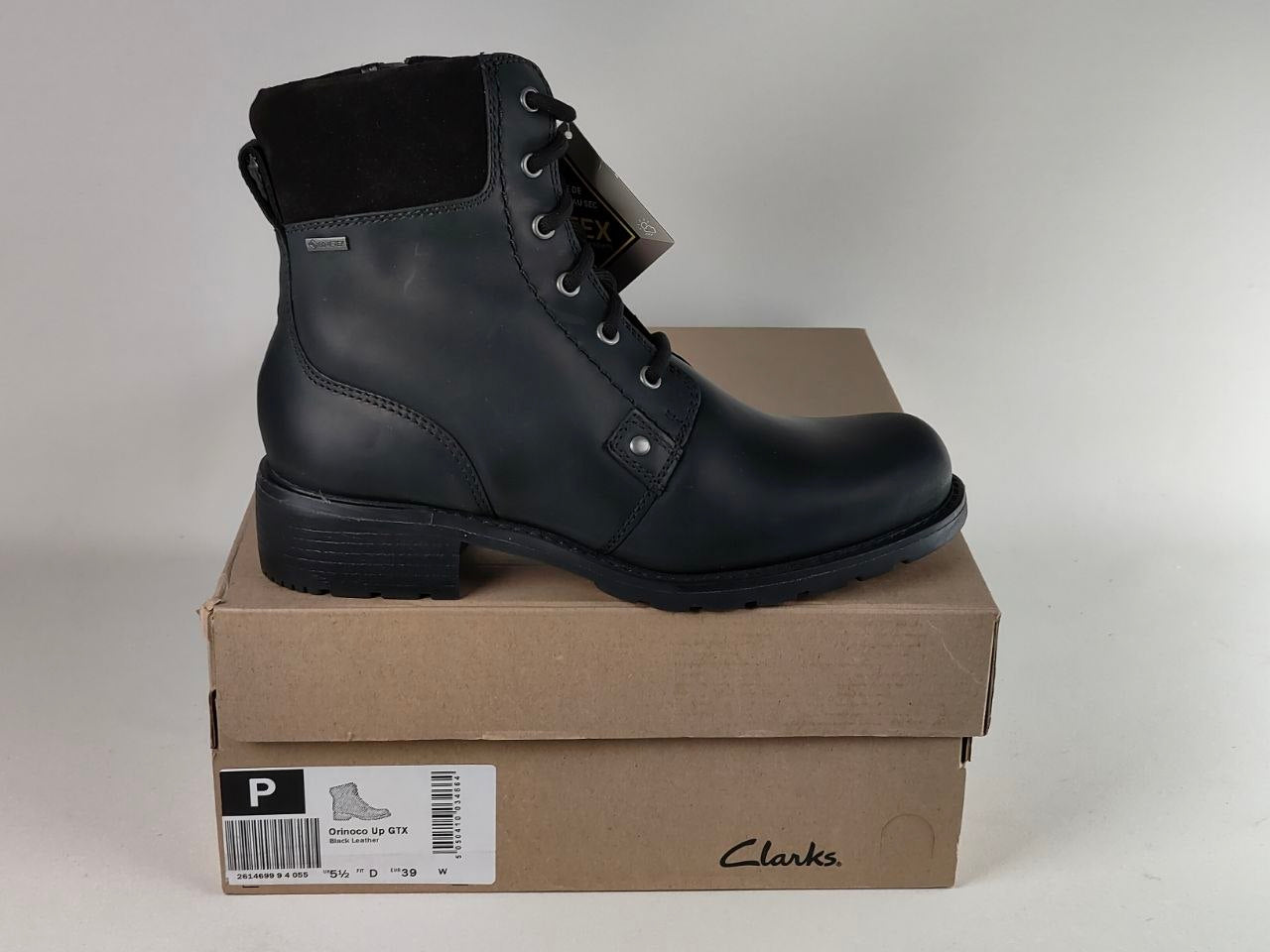 A black leather Clarks ankle boot with laces, displayed on top of its brown cardboard shoebox against a neutral light gray background.