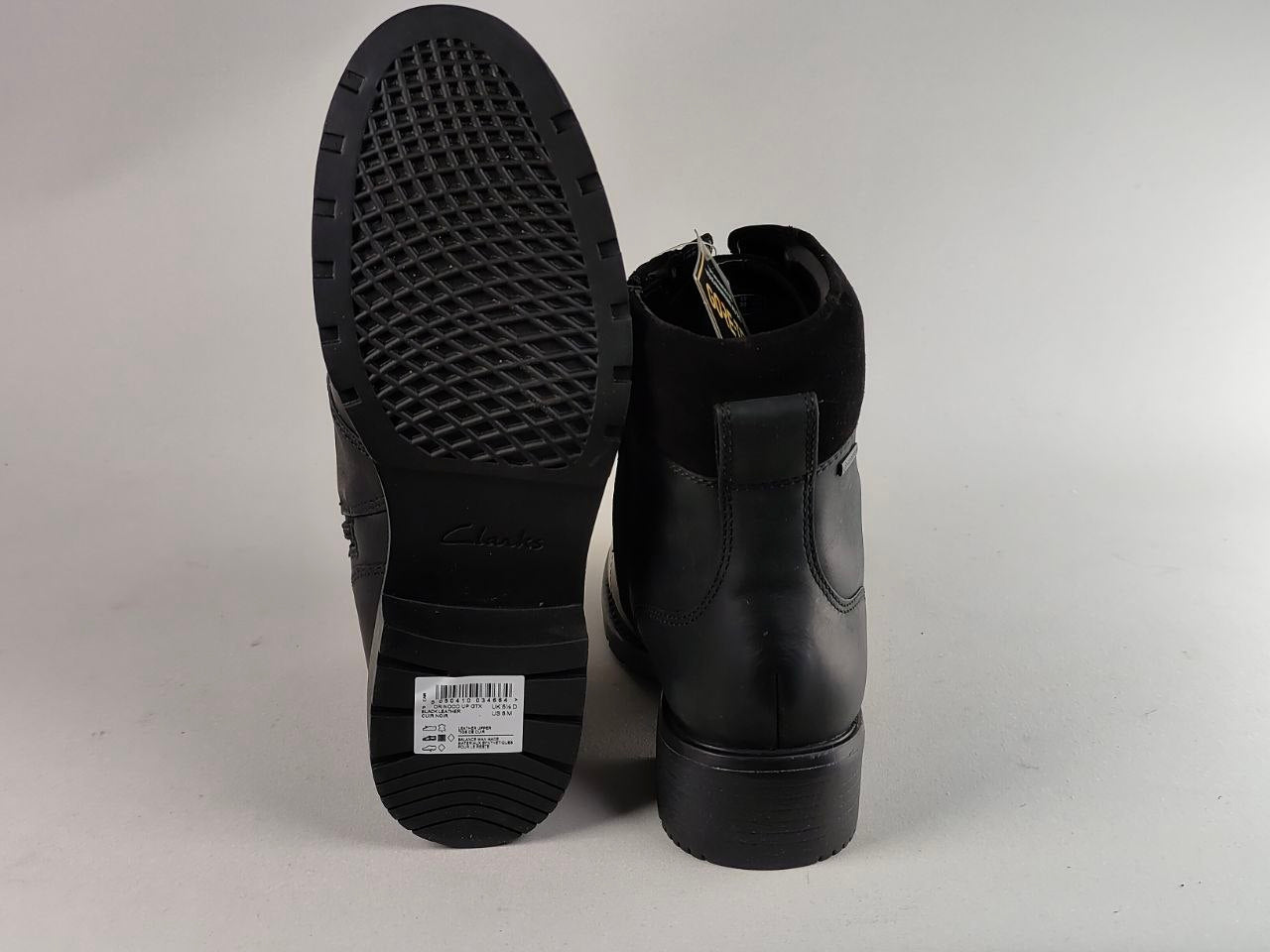 A pair of black ankle boots shown from the back, with one boot standing upright to display the side profile and the other tilted to show the textured black outsole. A white product tag is visible on the sole.