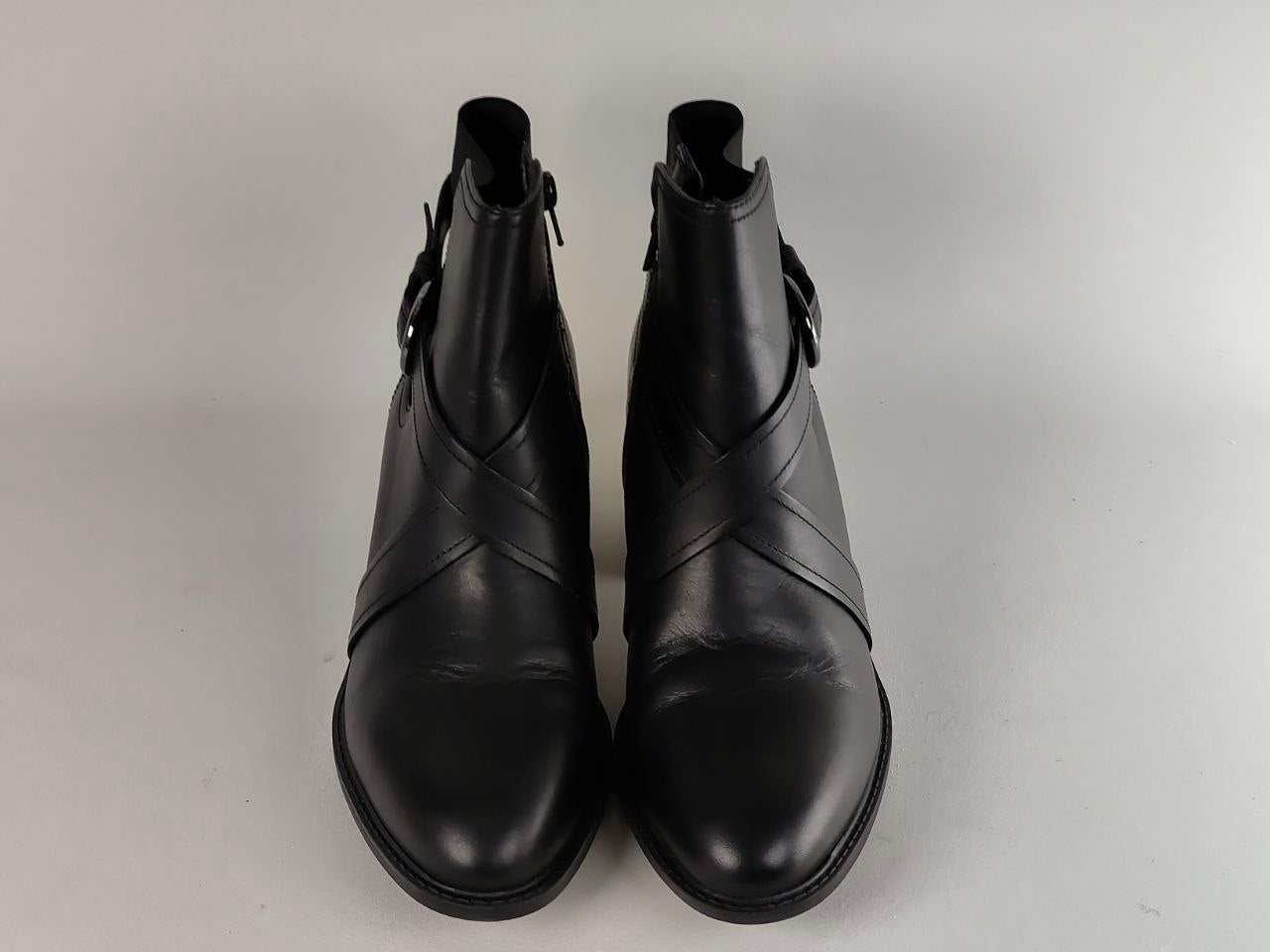 A pair of black leather ankle boots with criss-cross strap detailing across the front, photographed from a top-down angle on a light gray background.