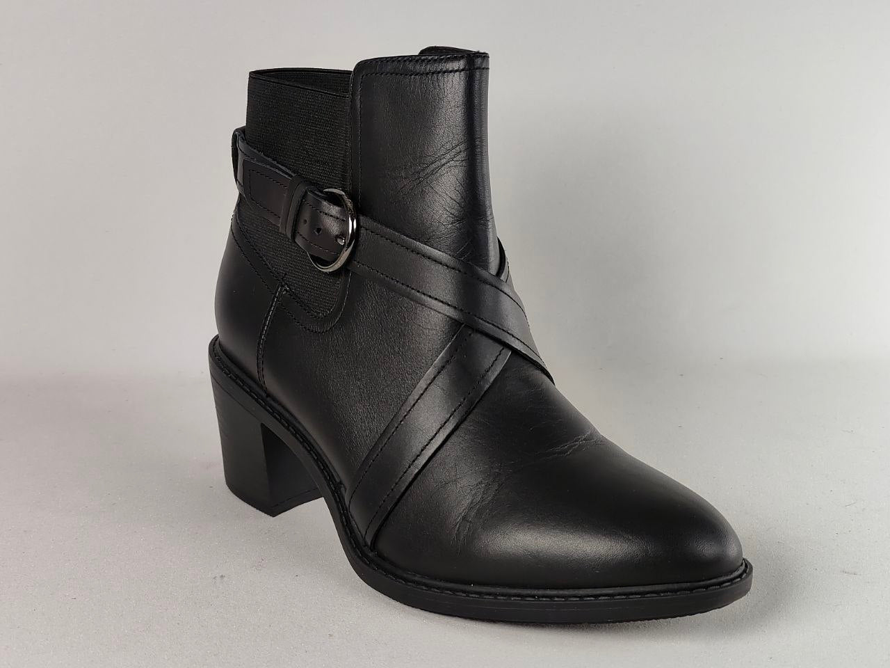 A black leather ankle boot with a block heel, featuring diagonal straps across the instep and a side buckle detail, shown against a neutral light gray background.