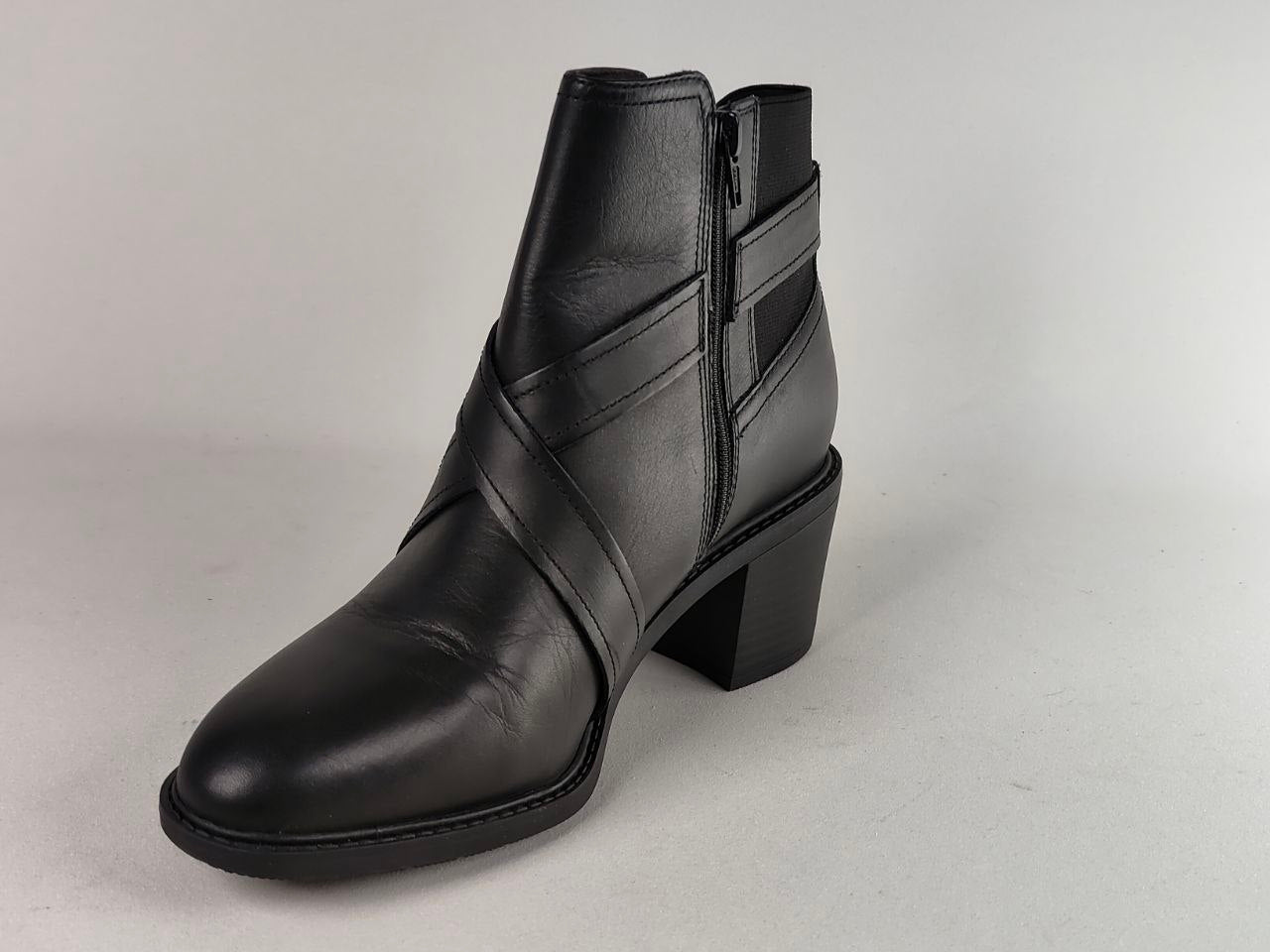A black leather ankle boot with a block heel, side zipper, and crisscross strap details on the side, shown against a neutral light gray background.