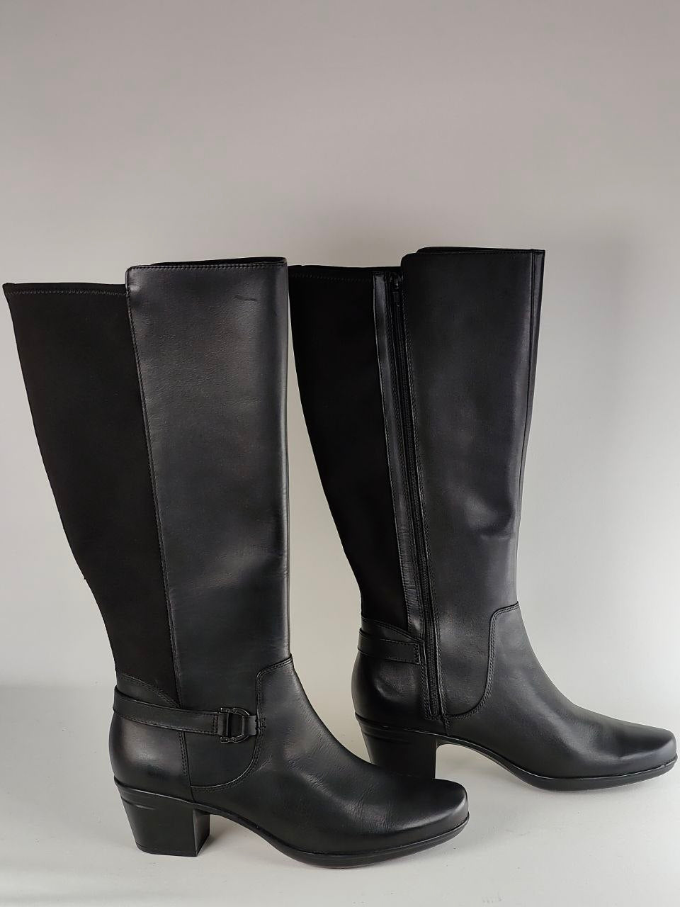 A pair of black knee-high leather boots with side zippers and a buckle strap detail on the front boot, shown against a neutral gray background.