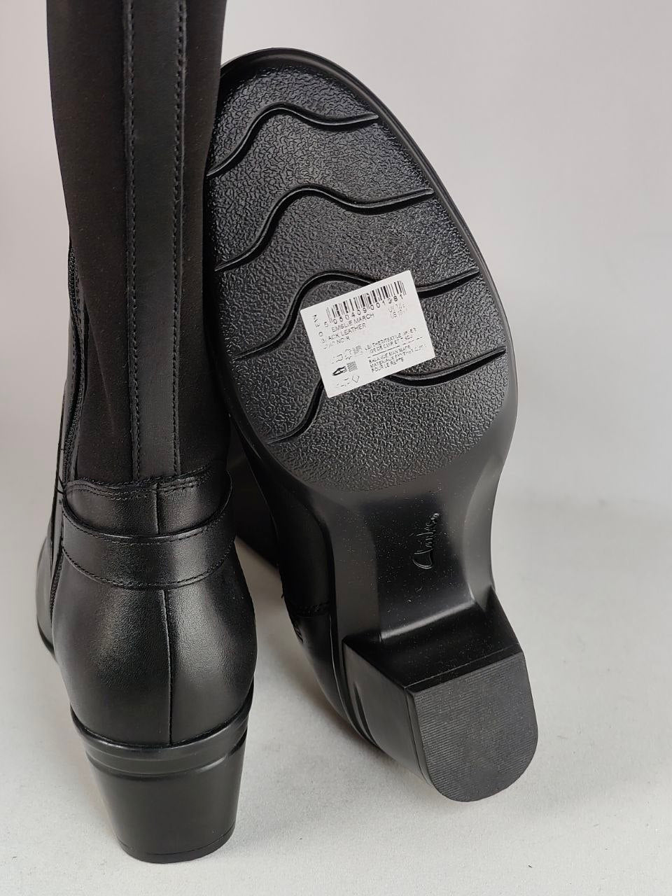 A pair of black knee-high boots shown from the back, with one boot standing upright and the other tilted to display the textured black rubber sole. A white product tag is visible on the sole.