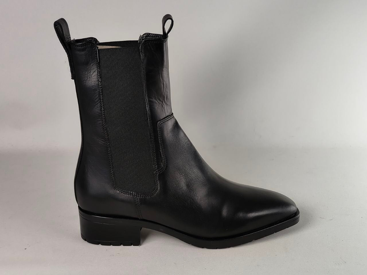 A black leather Chelsea-style ankle boot with a pointed toe, low block heel, and black elastic side gore, photographed against a neutral light gray background.