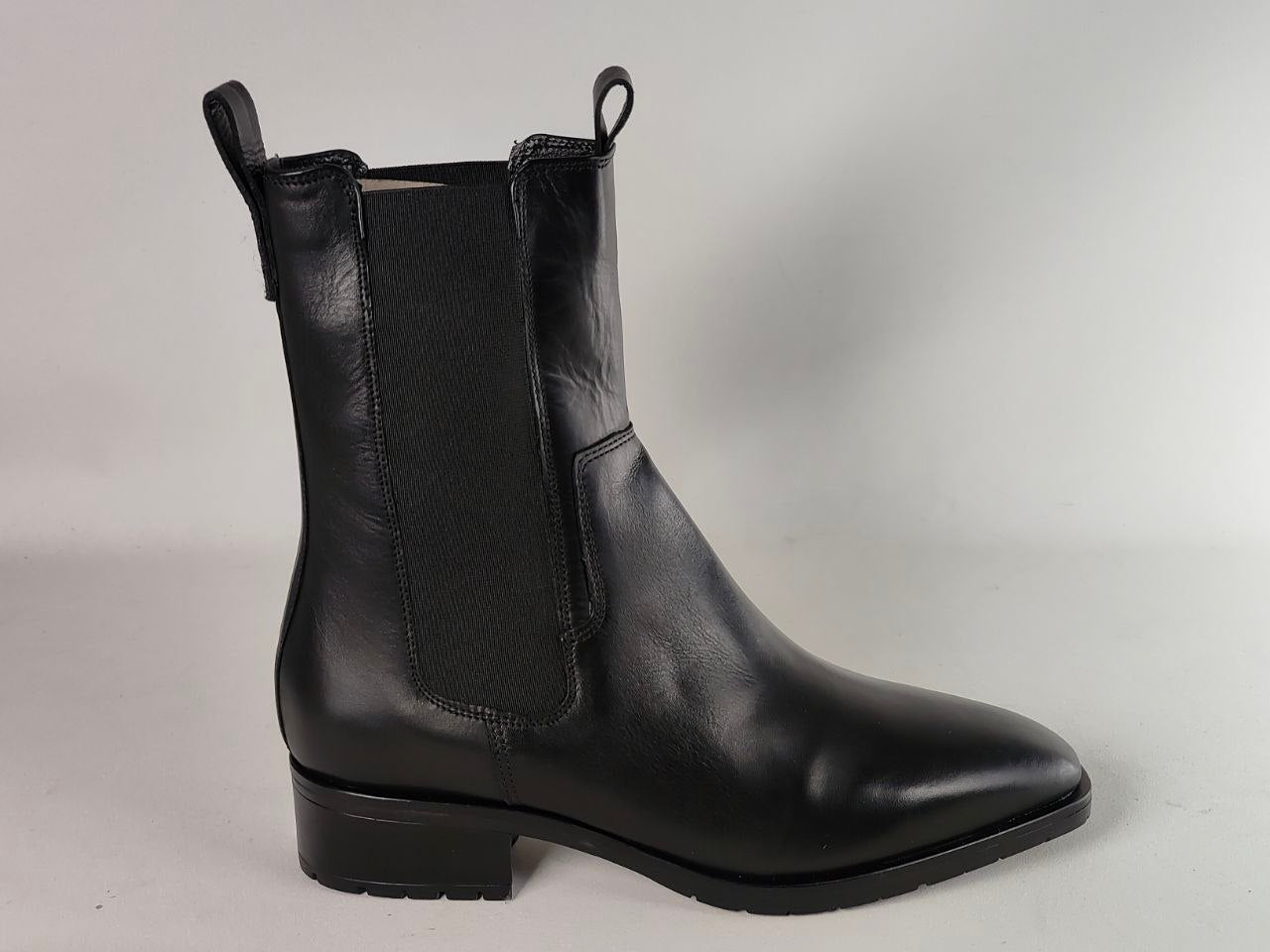 A black leather Chelsea-style ankle boot with a black elastic side panel, pull loops at the top, and a low block heel, shown from a side profile on a light gray background.