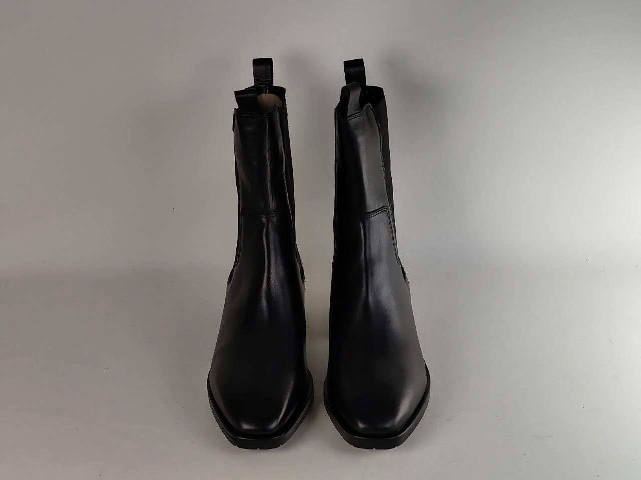 A pair of black leather Chelsea boots with side gussets and pull tabs, shown from the front against a neutral gray background.