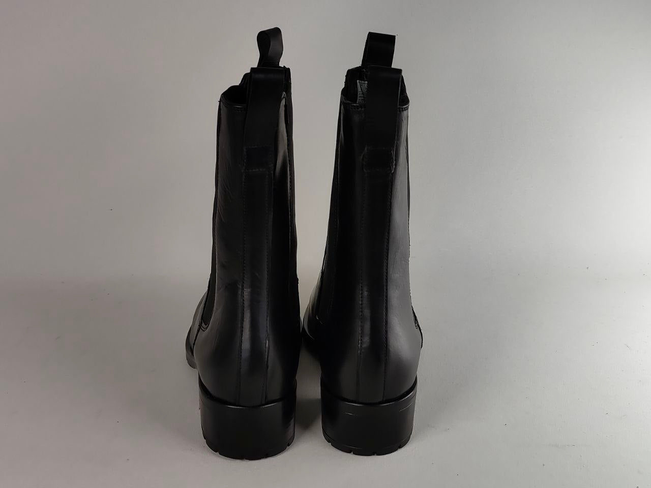 A pair of black leather ankle boots shown from the back, featuring a low block heel and pull tabs at the top, set against a plain light gray background.