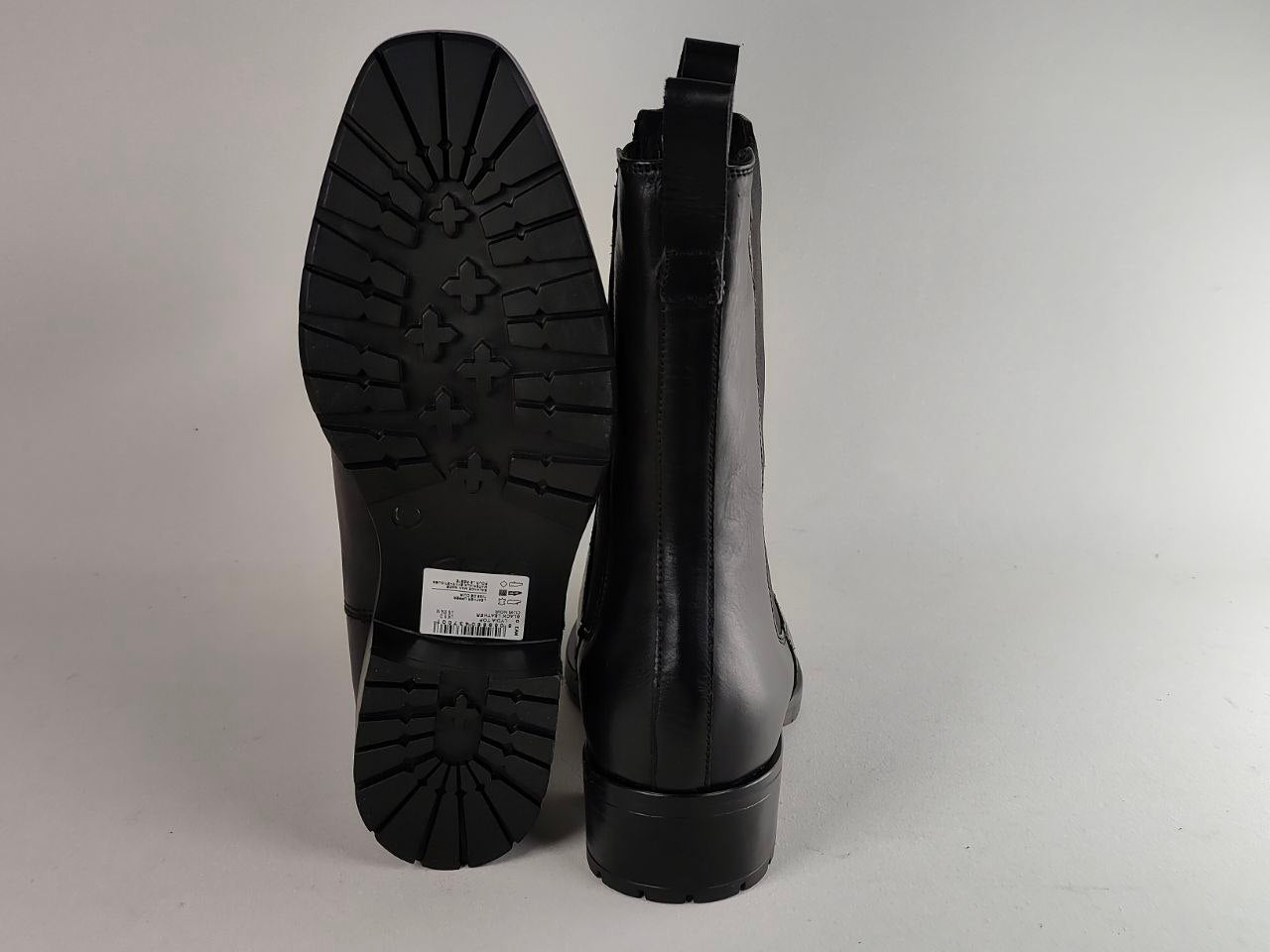 A pair of black leather ankle boots shown from the back, with one boot standing upright to display the heel and sole and the other leaning against it, on a light gray background.