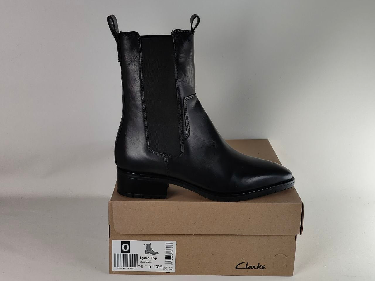 A black leather Chelsea boot with a low block heel, displayed on top of its brown Clarks shoebox against a neutral light gray background.