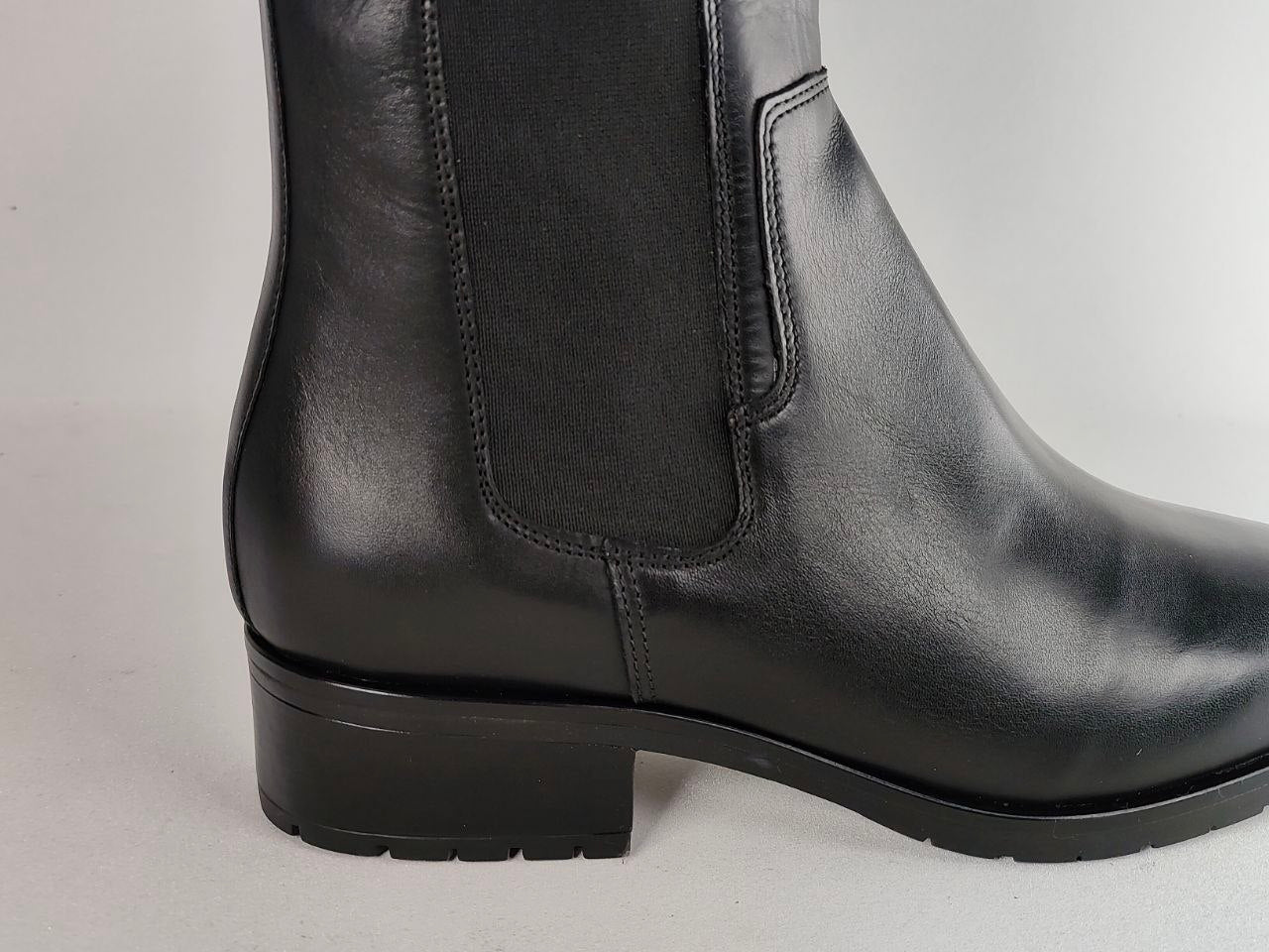 A close-up side view of a black leather ankle boot with a black elastic side panel and a low block heel, set against a plain light gray background.