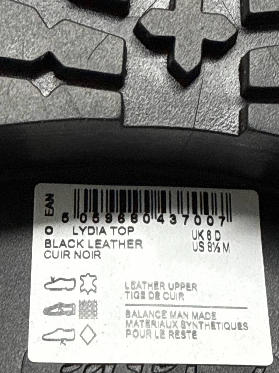 Close-up of a shoe sole label reading 'LYDIA TOP BLACK LEATHER' with a barcode and size information, set against a dark textured rubber background.