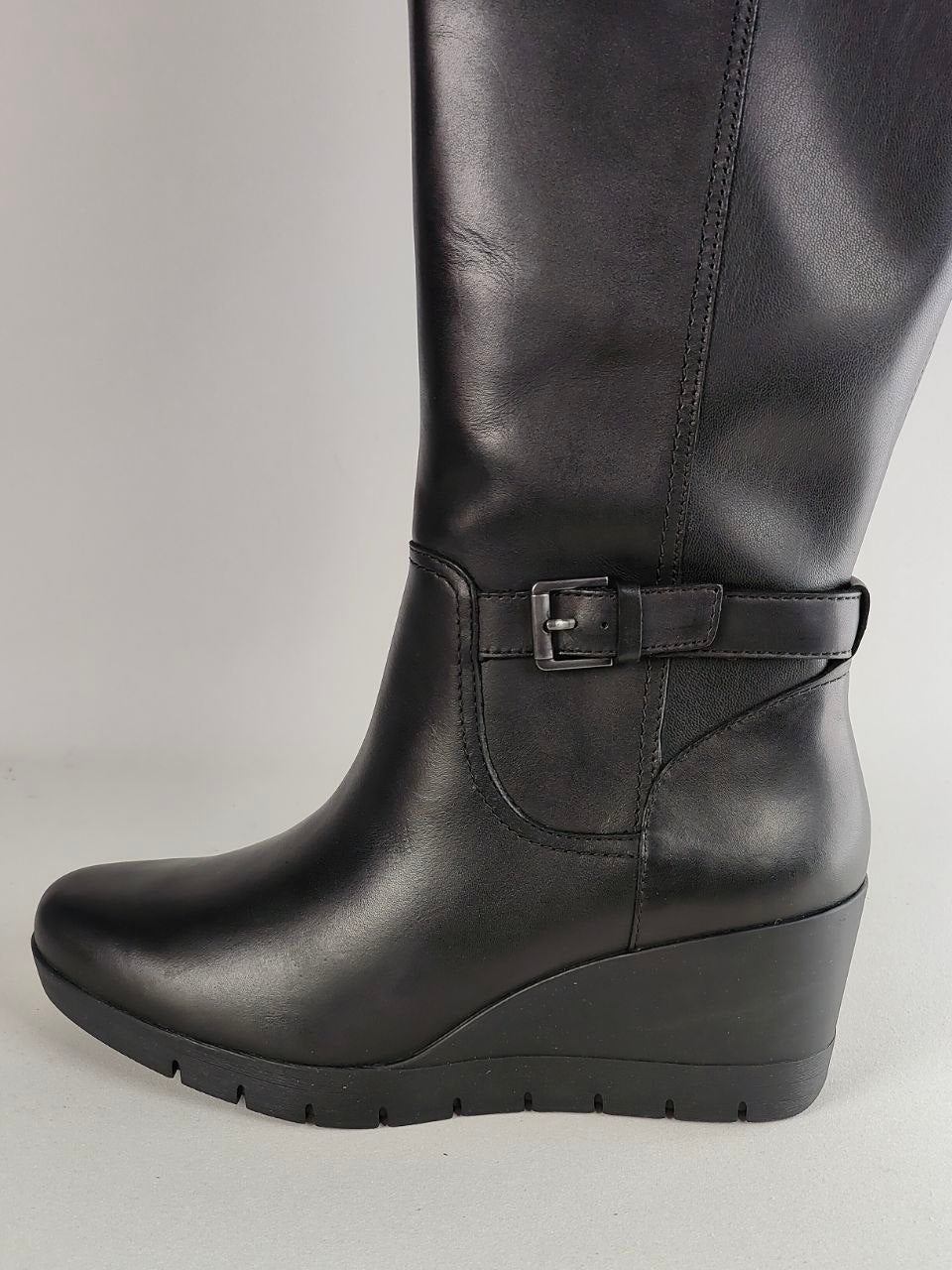 A black leather knee-high boot with a low wedge heel, a buckle strap detail around the ankle, and a rounded toe, shown against a neutral light gray background.