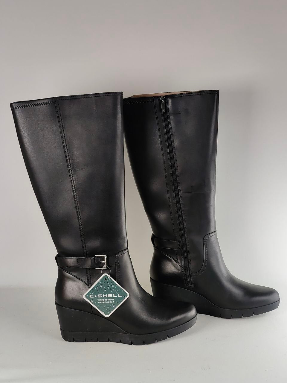 A pair of black leather knee-high riding boots with wedge heels, side zippers, and buckle straps, shown against a neutral gray background. A diamond-shaped C-SHELL brand tag is attached to the front boot.