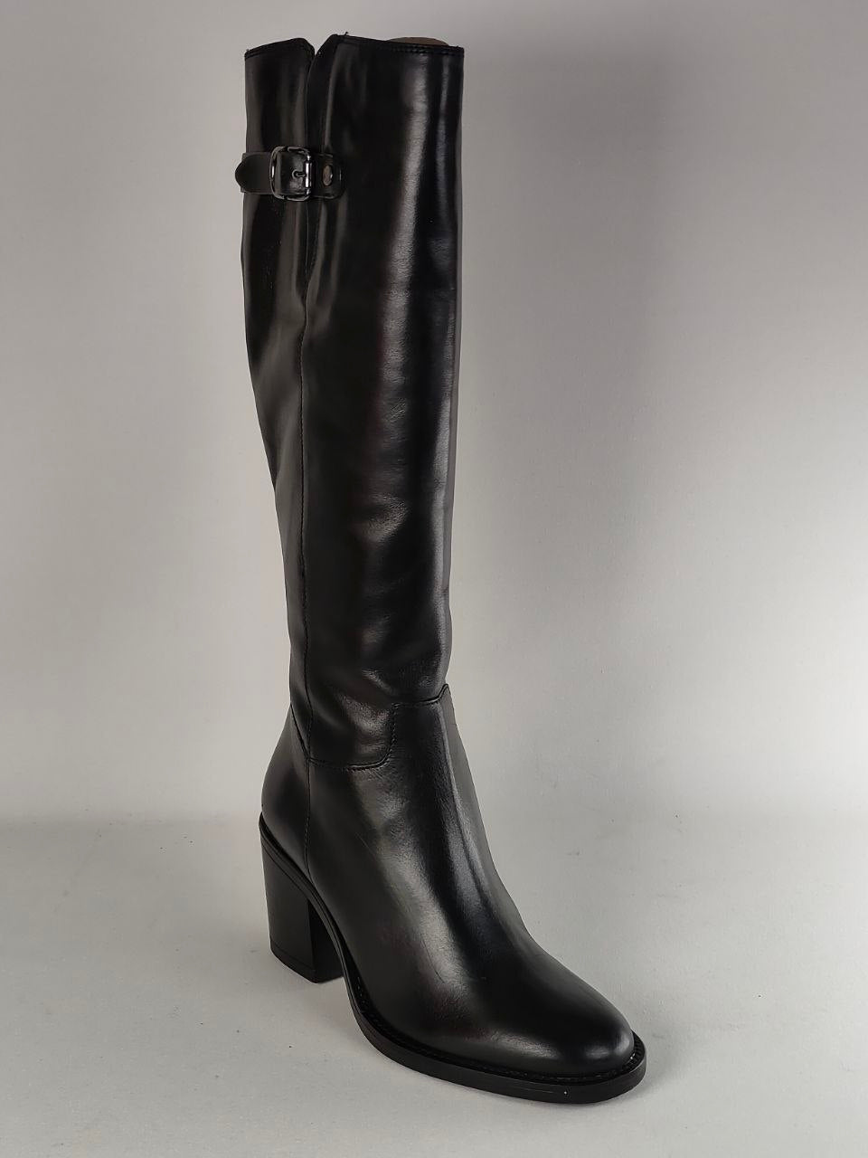 A single black leather knee-high boot with a block heel and a decorative buckle detail near the top, photographed against a neutral light gray background.