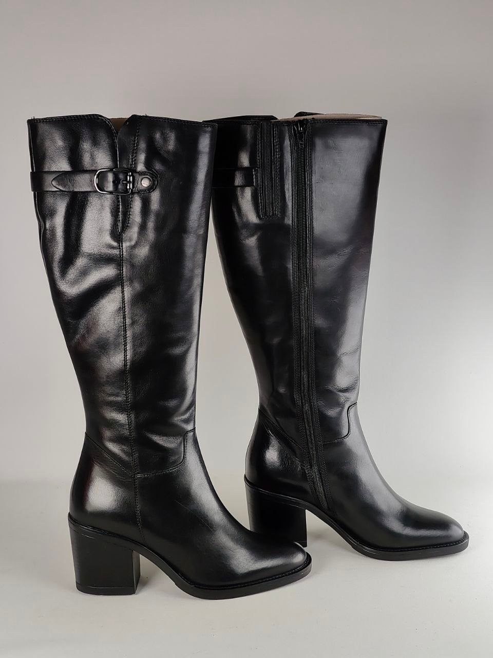 A pair of black leather knee-high boots with block heels, shown against a neutral gray background. One boot is standing upright, displaying its side zipper and decorative strap detail, while the other is angled to show the front profile.
