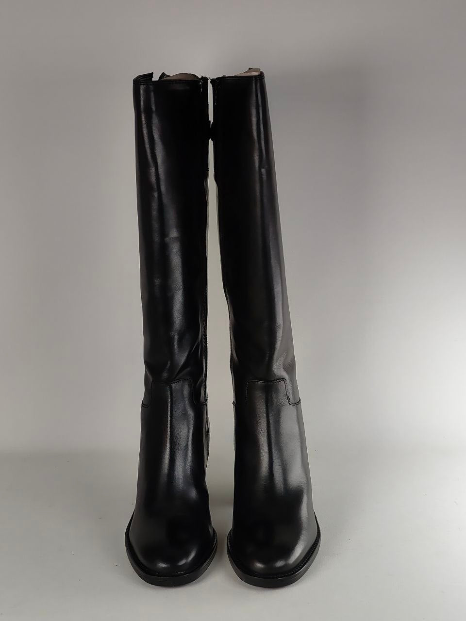 A pair of black leather knee-high boots with a pointed toe and low block heel, photographed against a neutral gray background.