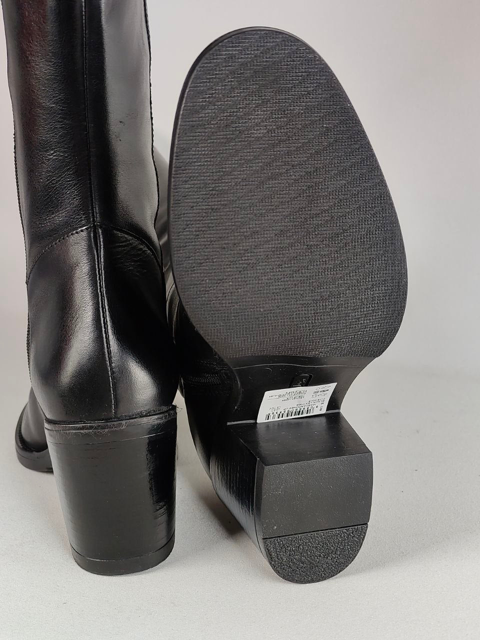 A pair of black leather boots shown from the back, featuring a high block heel and a textured black rubber outsole, photographed against a neutral light gray background.