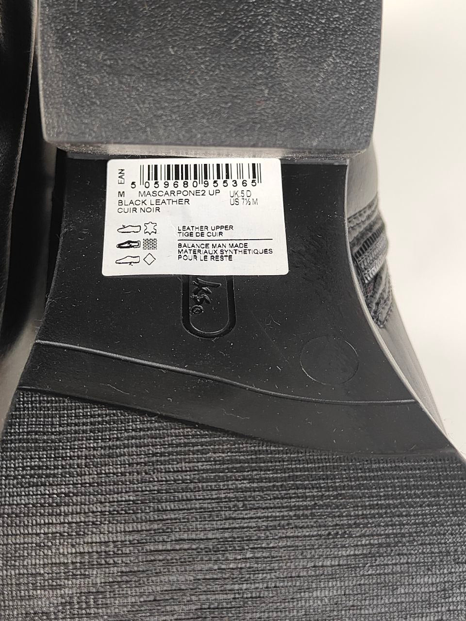 Close-up of the heel area of a black leather boot, showing a white barcode label with product details and a textured black sole.
