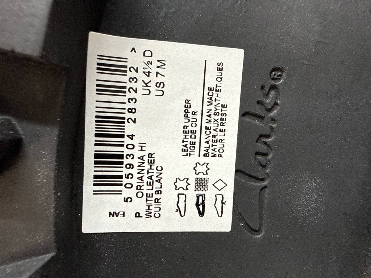 A close-up of a white product label with a barcode and printed text on a dark gray or black plastic surface. The label includes text such as 'ORIANNA HI', 'PUDDLE LEATHER', 'OUR BLANC', and material icons, with a stylized script logo that appears to read 'Clark'.