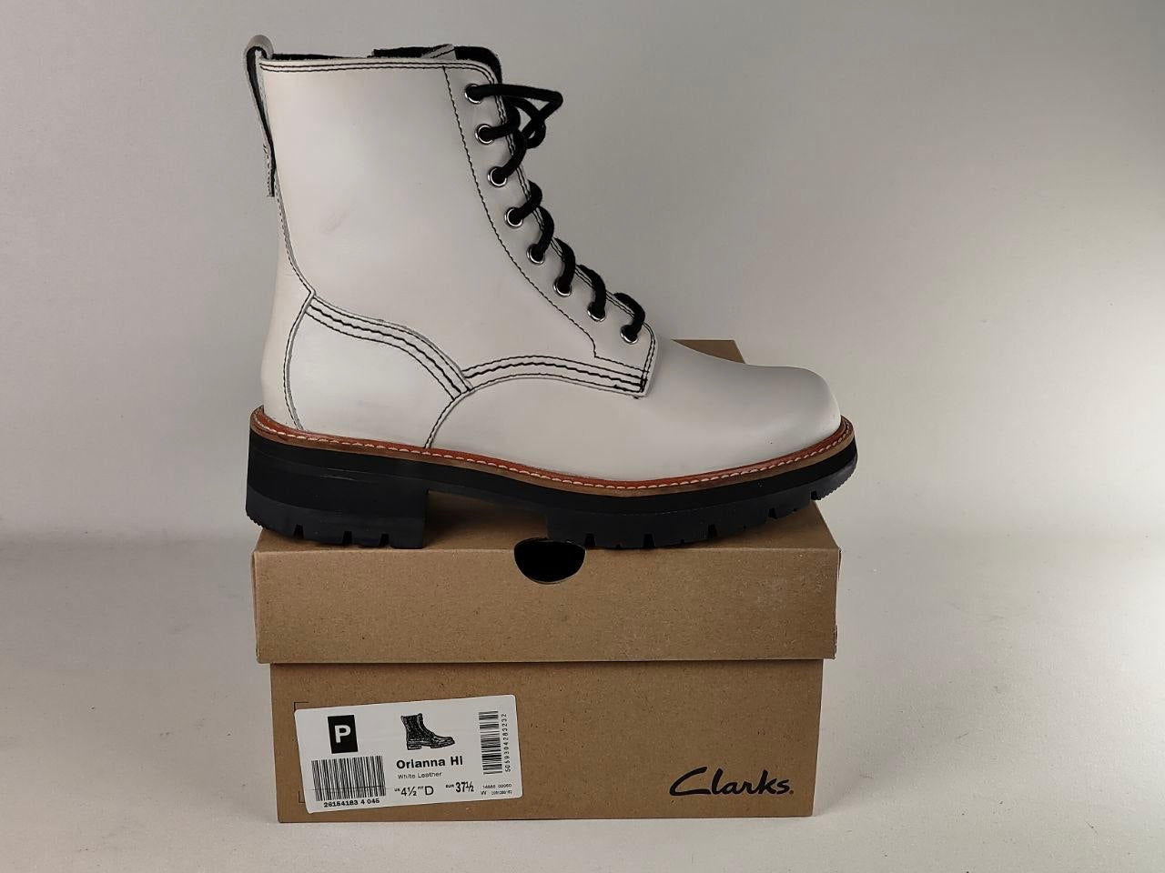 A white leather lace-up boot with black laces and a chunky black sole, displayed on top of a brown Clarks shoebox against a light gray background.