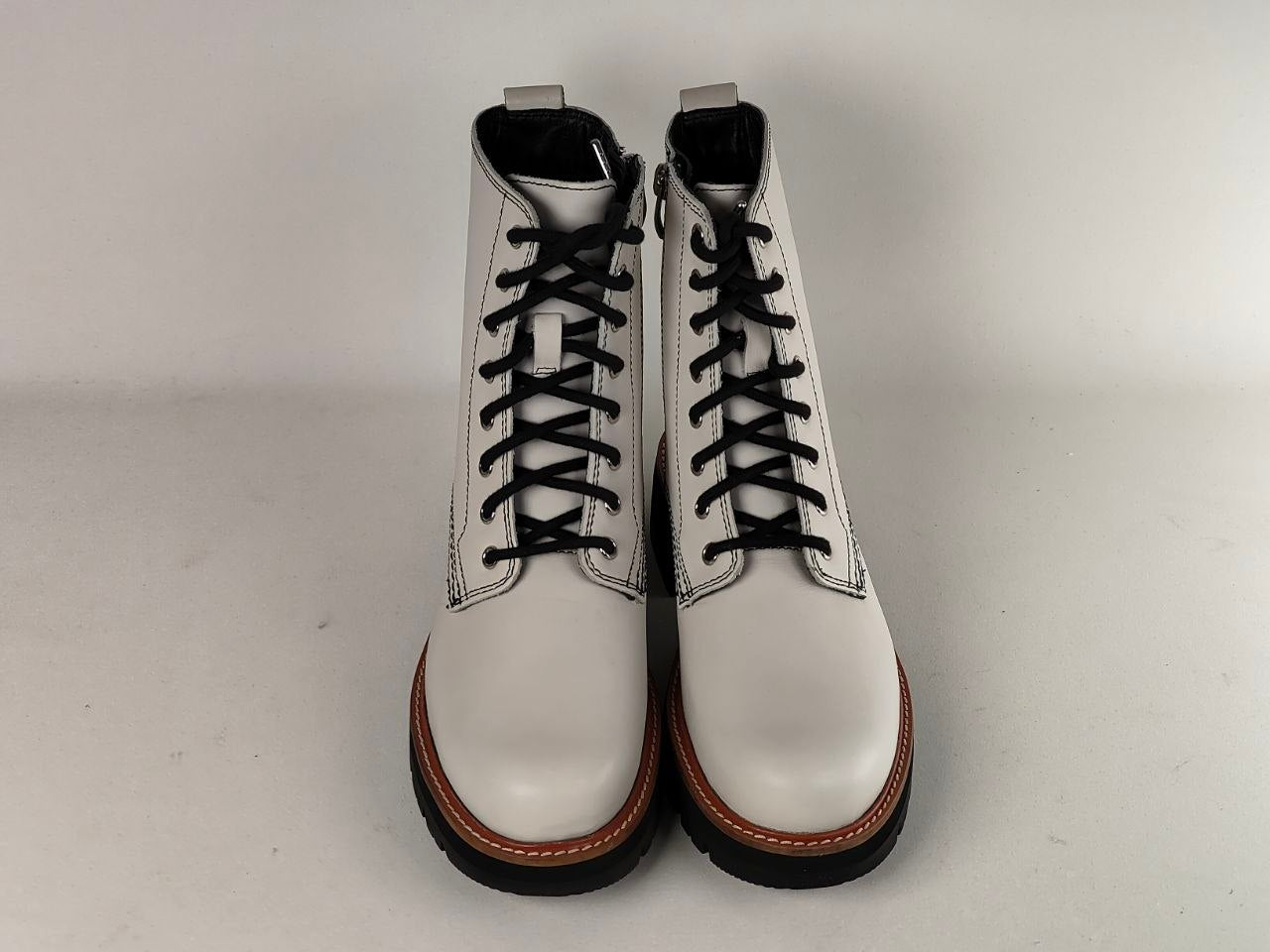 A pair of white leather lace-up ankle boots with black laces, black soles, and brown stitching around the sole edge, photographed against a plain light gray background.
