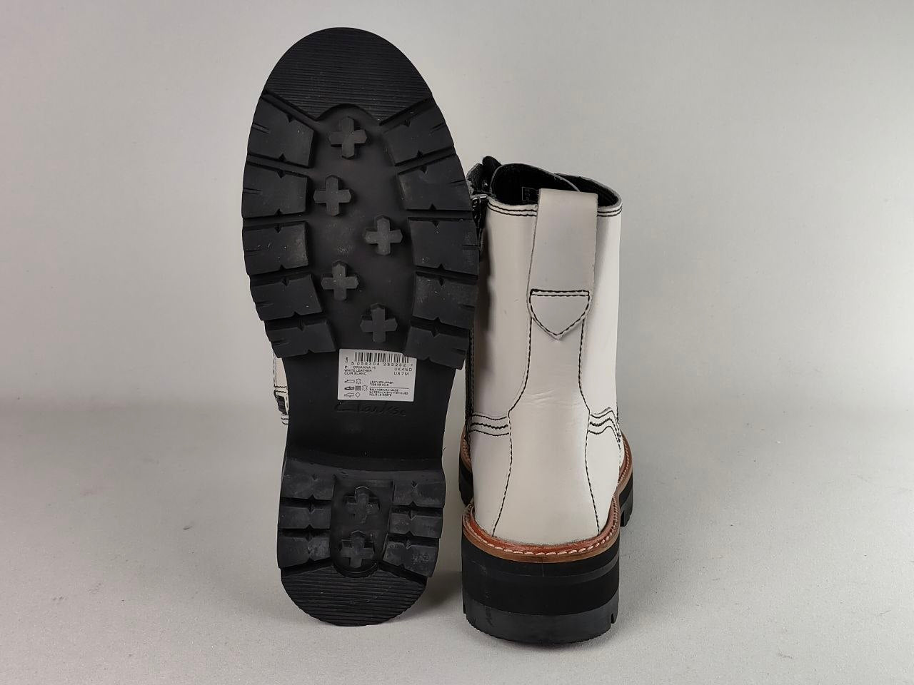 A pair of white leather ankle boots shown from the back, with one boot standing upright to display the white leather upper, stitching, heel tab, and brown welt, and the other boot leaning on its black lug sole.