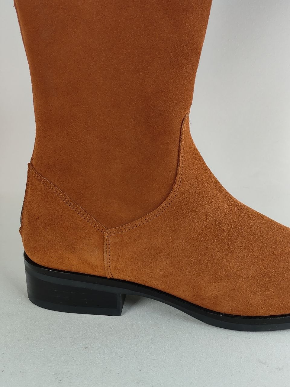 A close-up side view of a brown suede boot with a black block heel and sole, set against a plain light gray background.