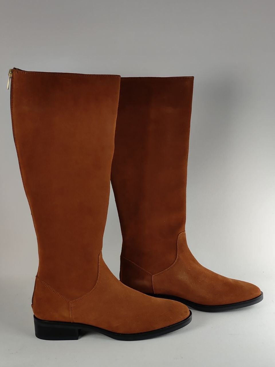 A pair of tall brown suede boots with black low block heels, shown against a neutral gray background.