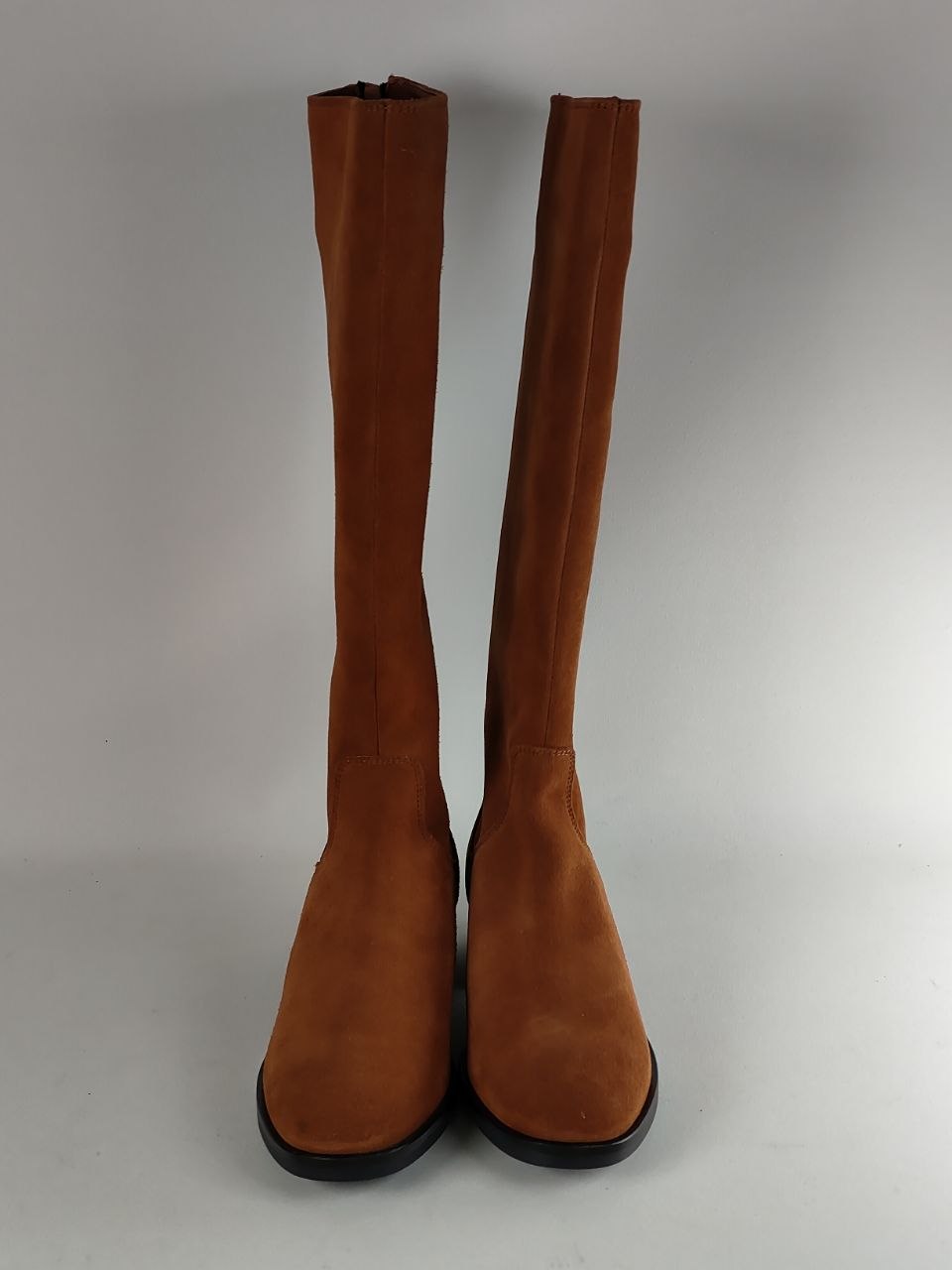 A pair of tall brown suede knee-high boots with black soles, photographed against a neutral gray studio background.