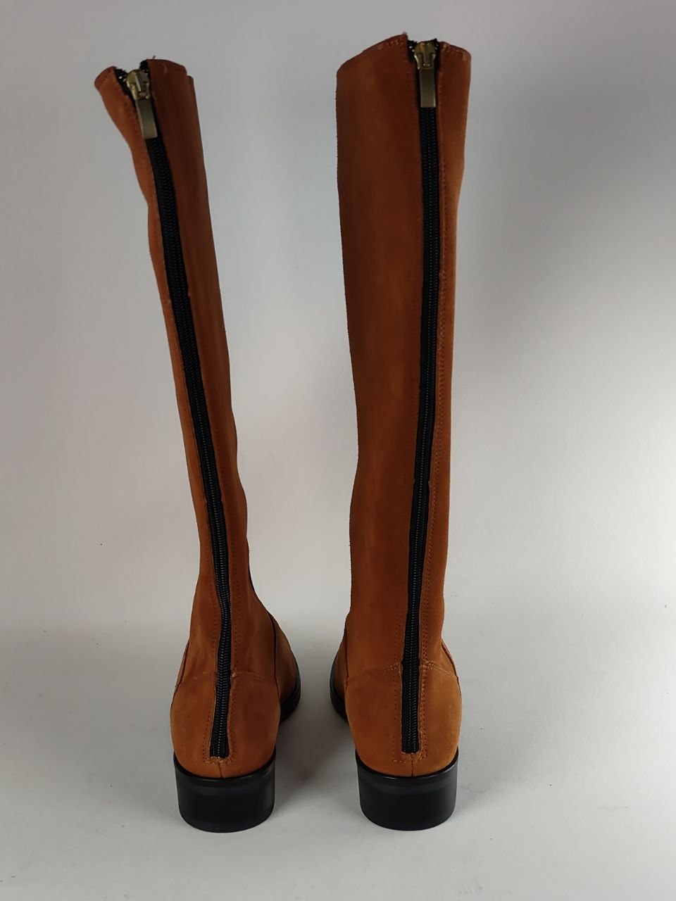 A pair of tall brown suede boots shown from the back, featuring black zippers running down the center and black soles, set against a plain light gray background.