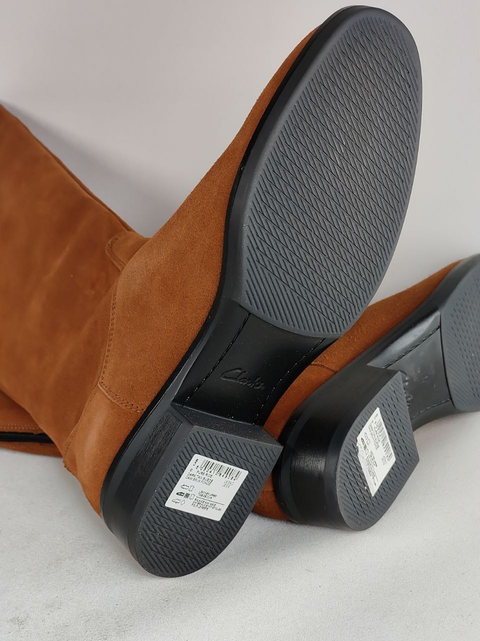 A pair of brown suede boots shown from the sole, displaying the black treaded outsole and heel area with product labels visible.
