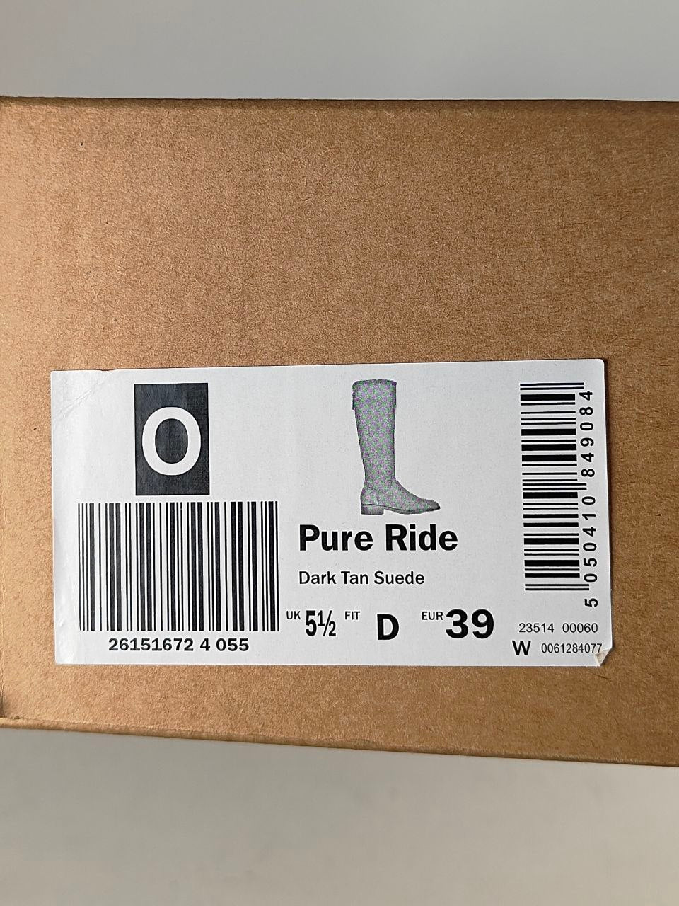 A close-up of a brown cardboard shoebox with a white product label. The label shows the brand 'Pure Ride', the style 'Dark Tan Suede', a silhouette of a tall boot, and barcode details.