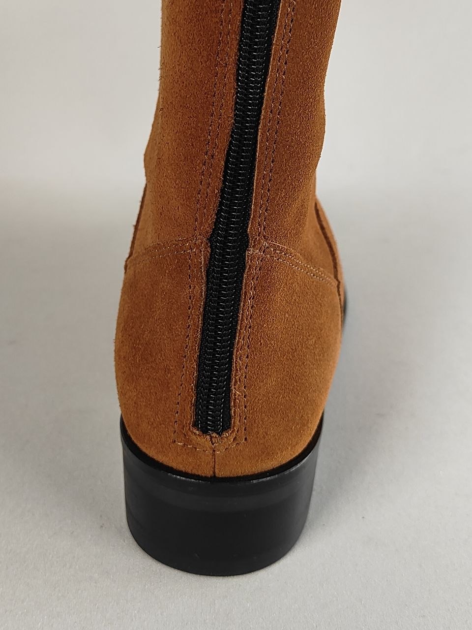 Close-up rear view of a brown suede ankle boot with a black zipper running vertically and a black block heel, shown on a light gray background.