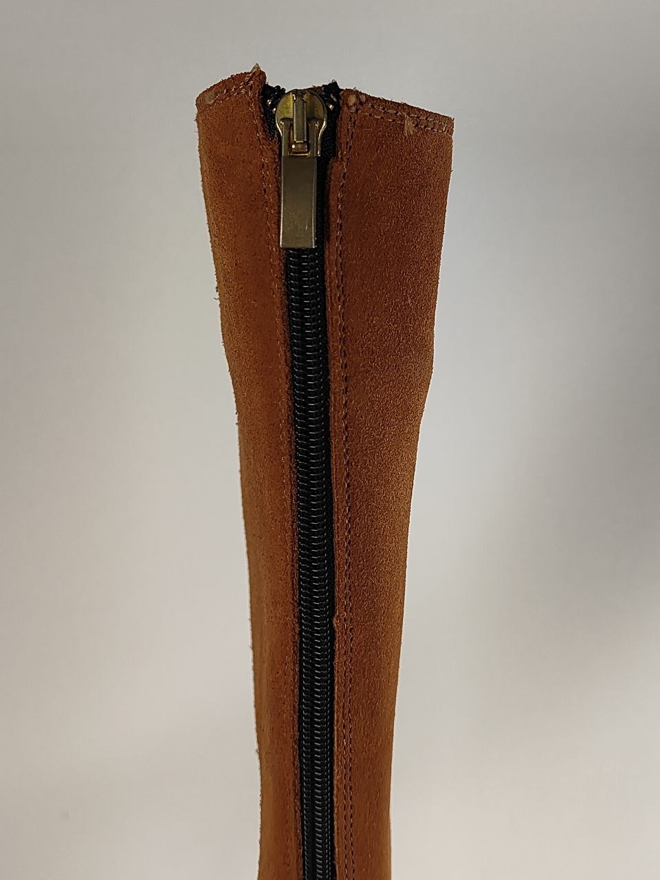 A close-up of a brown suede or nubuck boot shaft with a black center zipper and a brass-toned metal pull tab, set against a plain light gray background.