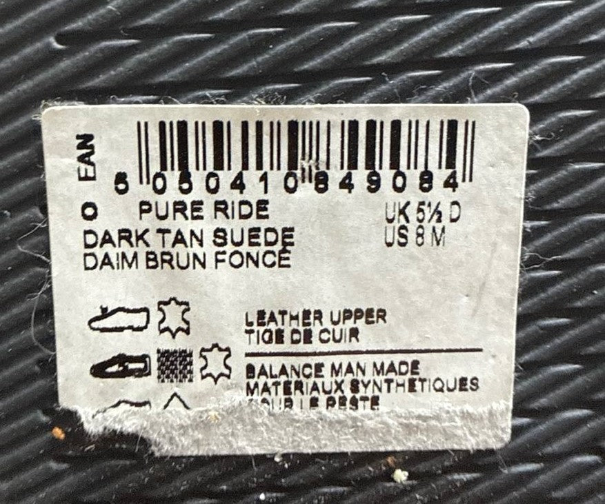 A close-up of a white paper label with a barcode and printed text, including 'PURE RIDE', 'DARK TAN SUEDE', and 'DAIM BRUN FONCE', placed on a dark textured surface.