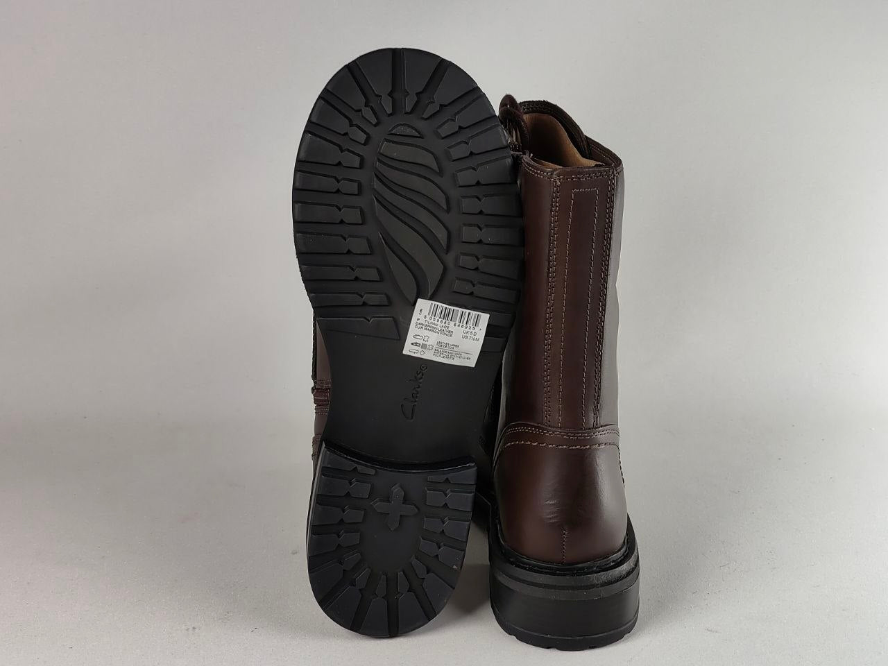 A pair of dark brown leather ankle boots shown from the back, with one boot standing upright to display the side and heel, and the other lying flat to show the black treaded outsole. A white size tag is visible on the sole.
